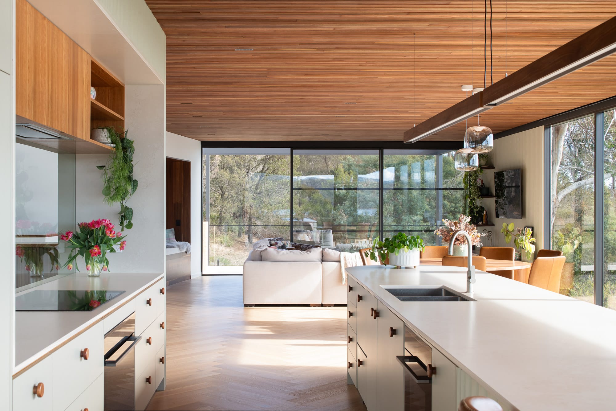 Mortlock Timber Series: The Milldam. Photography by Anjie Blair. Open plan kitchen and living space with white cabinetry and lounge, timber clad ceilings and timber herringbone floors. Floor-to-ceiling windows overlooking bushland. 