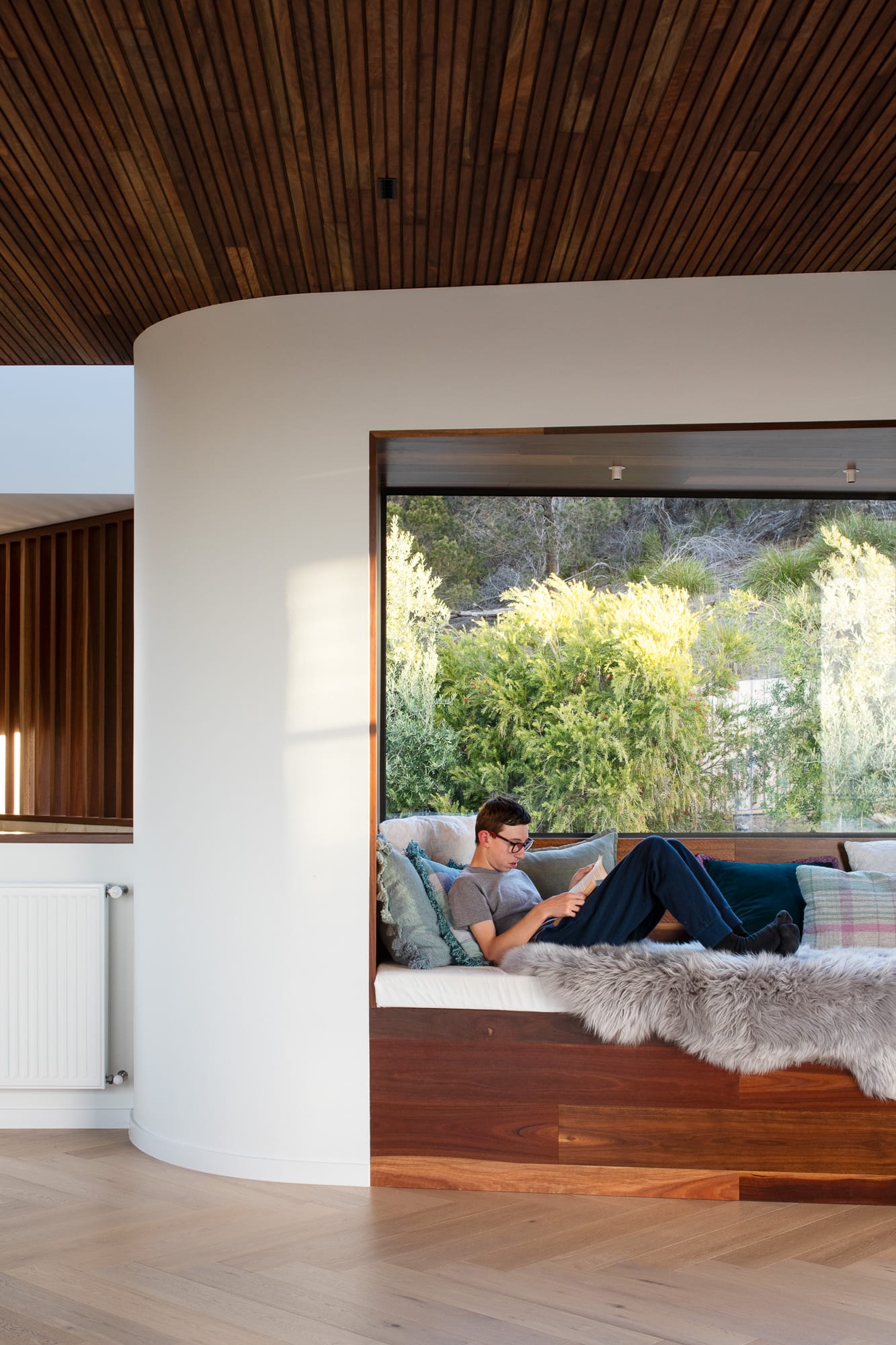 Mortlock Timber Series: The Milldam. Photography by Anjie Blair. Boy reading in nook with white curve exterior walls, timber base and large window overlooking garden. 