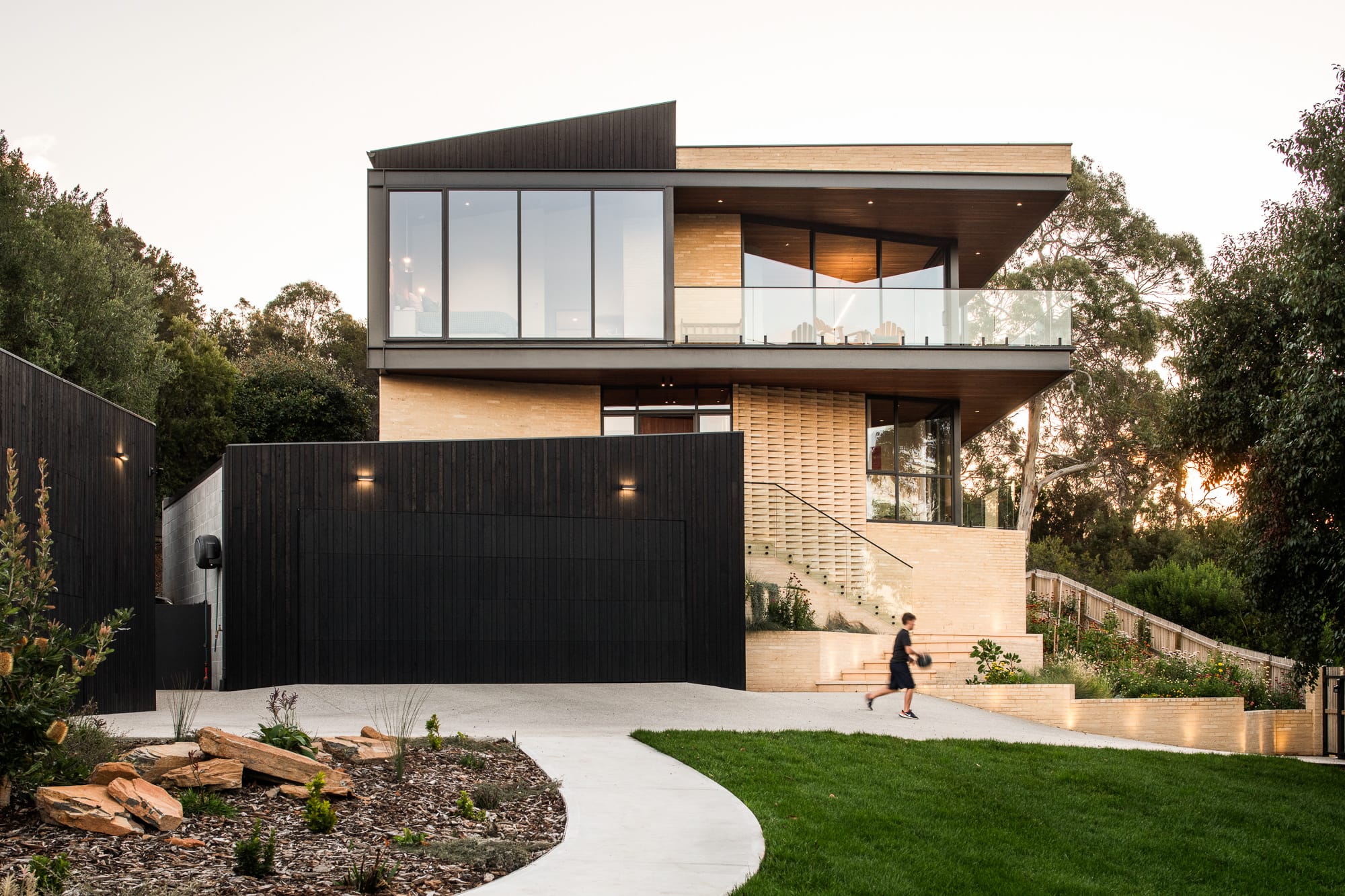 Mortlock Timber Series: The Milldam. Photography by Anjie Blair. Frontage of double storey residential home with black timber clad garage and winding pathway up to house. 