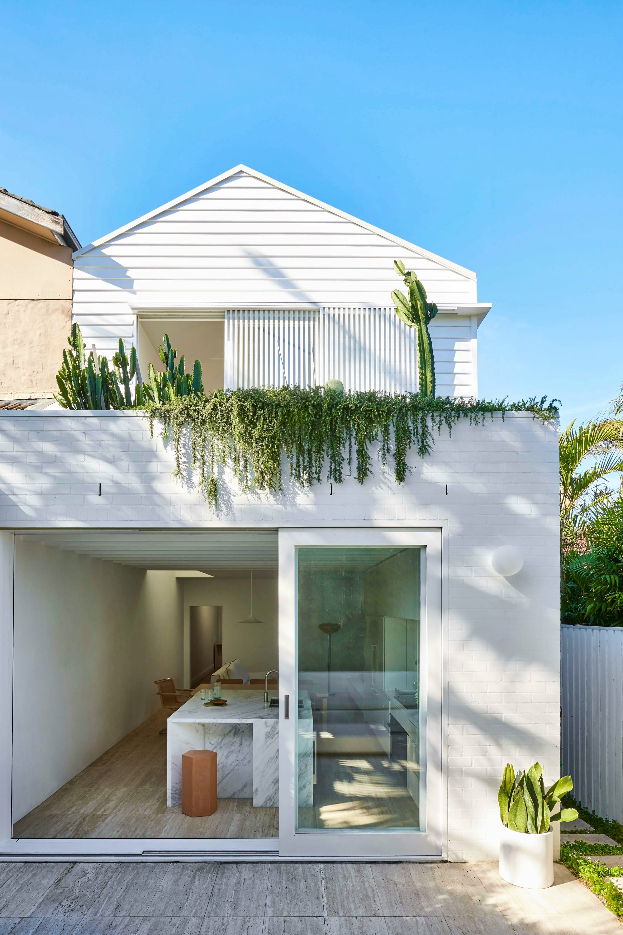 North Bondi House by Stdio Arkive. Photography by Pablo Veiga. Exterior facade of double storey residential property finished with white brick and white timber clad. Glass sliding door looking onto kitchen with marble island bench. 