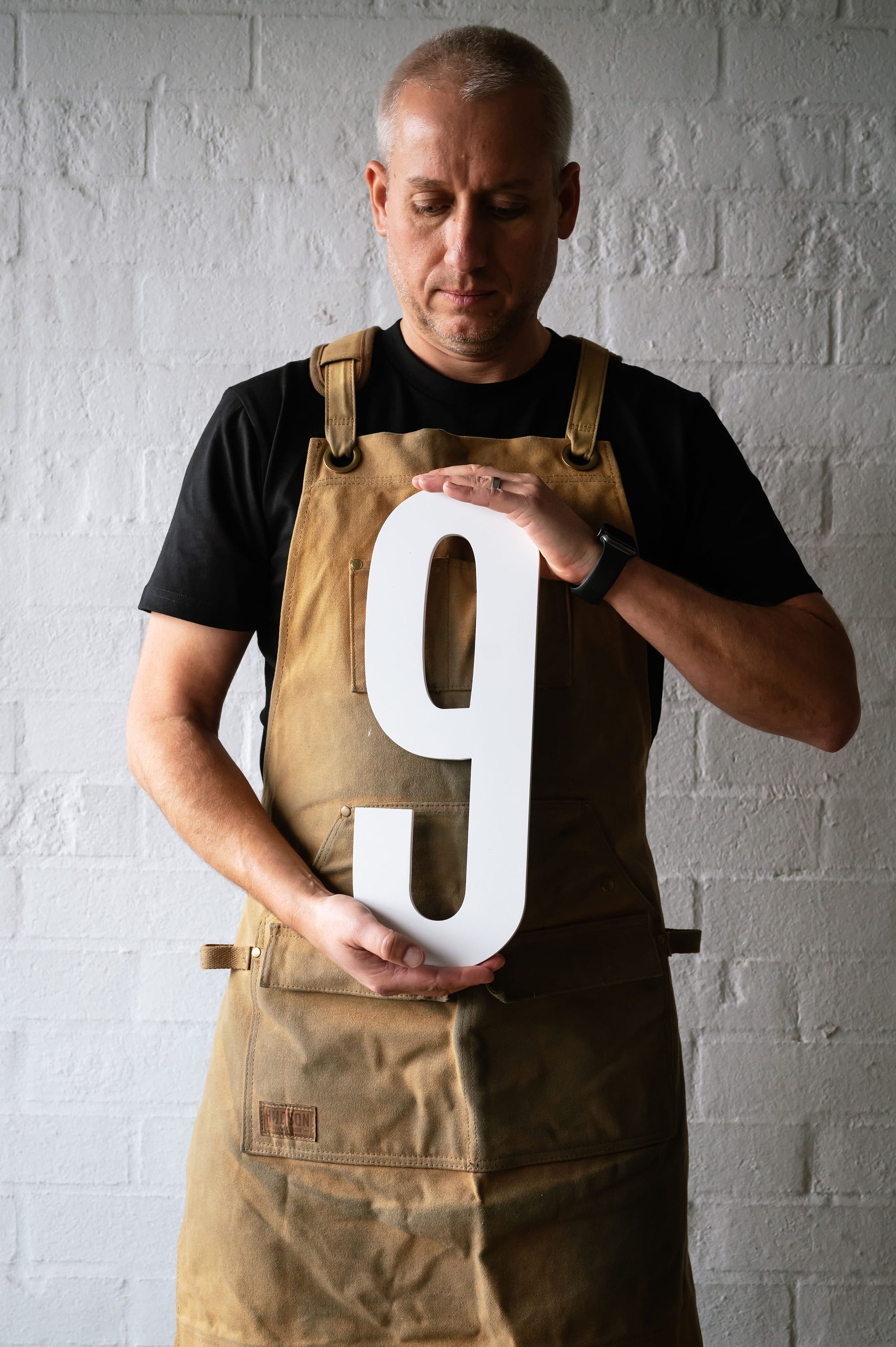 Peninsula House Numbers. Image copyright of Peninsula House Numbers. Man in black shirt and tan apron holding large white number 9 sign.