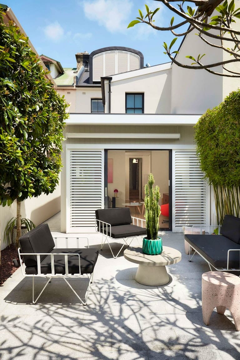 Redfern Reno by Blake Letnic Architects. Photography by Prue Ruscoe and Chloe Rayfield. Rear facade of double storey white home. White wire outdoor furniture with black cushions on grey tiled patio. Cactus plant on concrete coffee table. 