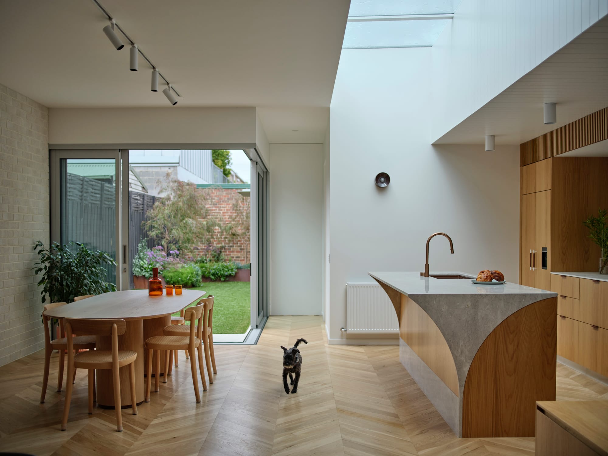ZigZag House by True Story. Photography by Dean Bradley. Kitchen and dining room with herringbone timber floors, timber kitchen counters and dining table. Opens onto grassed courtyard.