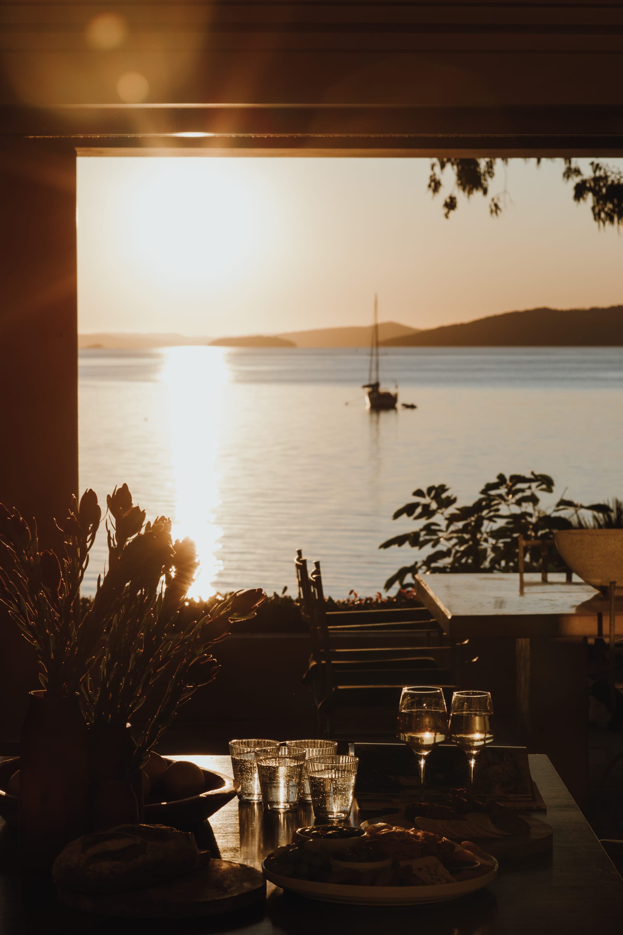 Amarna Luxury Beach Resort. Photography by Will Salkeld. Blurred view of harbour with sail boat and hills in background. Dining venue in foreground with tables and chairs with wine and food. 