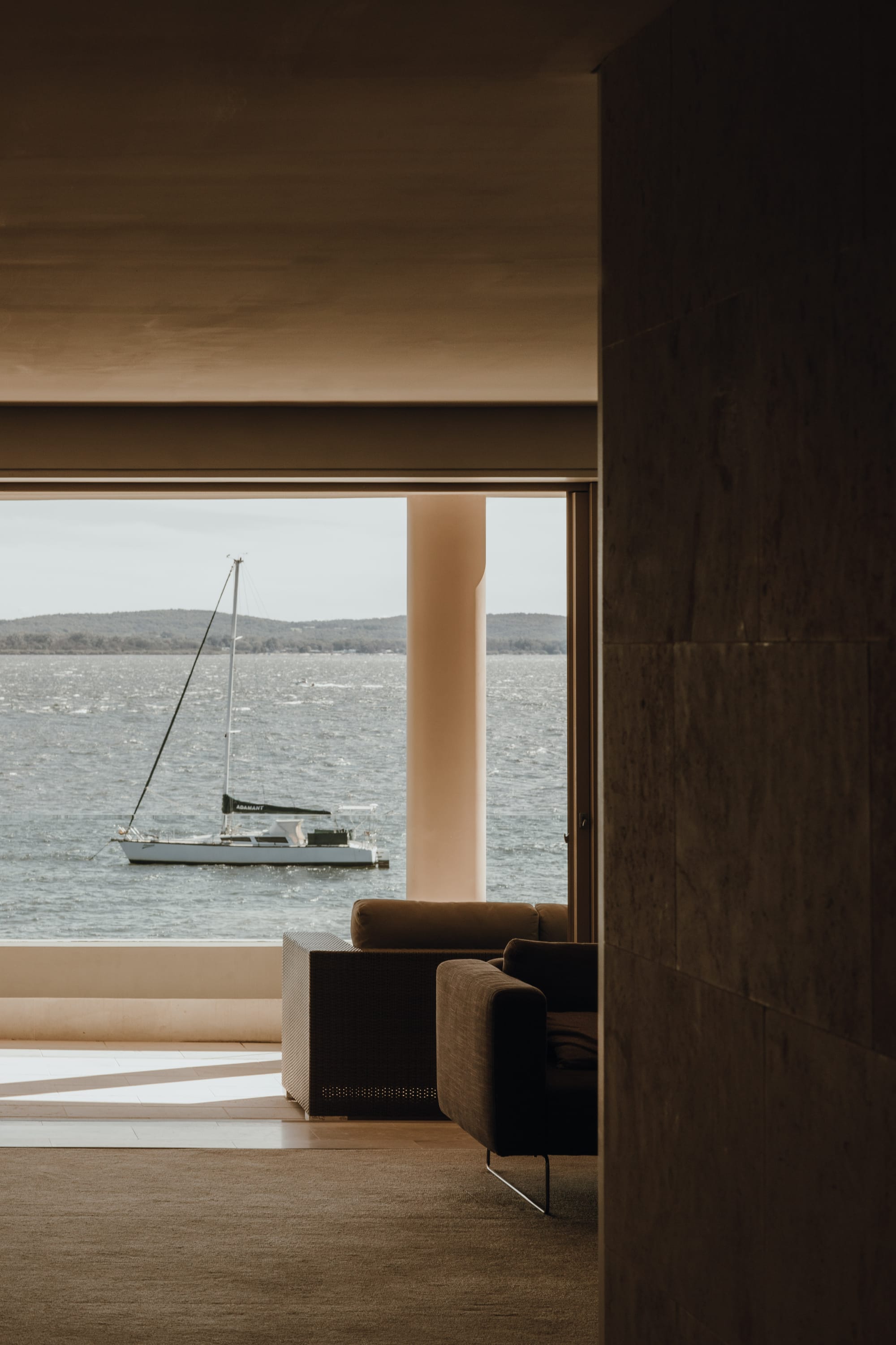 Amarna Luxury Beach Resort. Photography by Will Salkeld. Sailboat on open water framed through window with dark lounges in foreground.