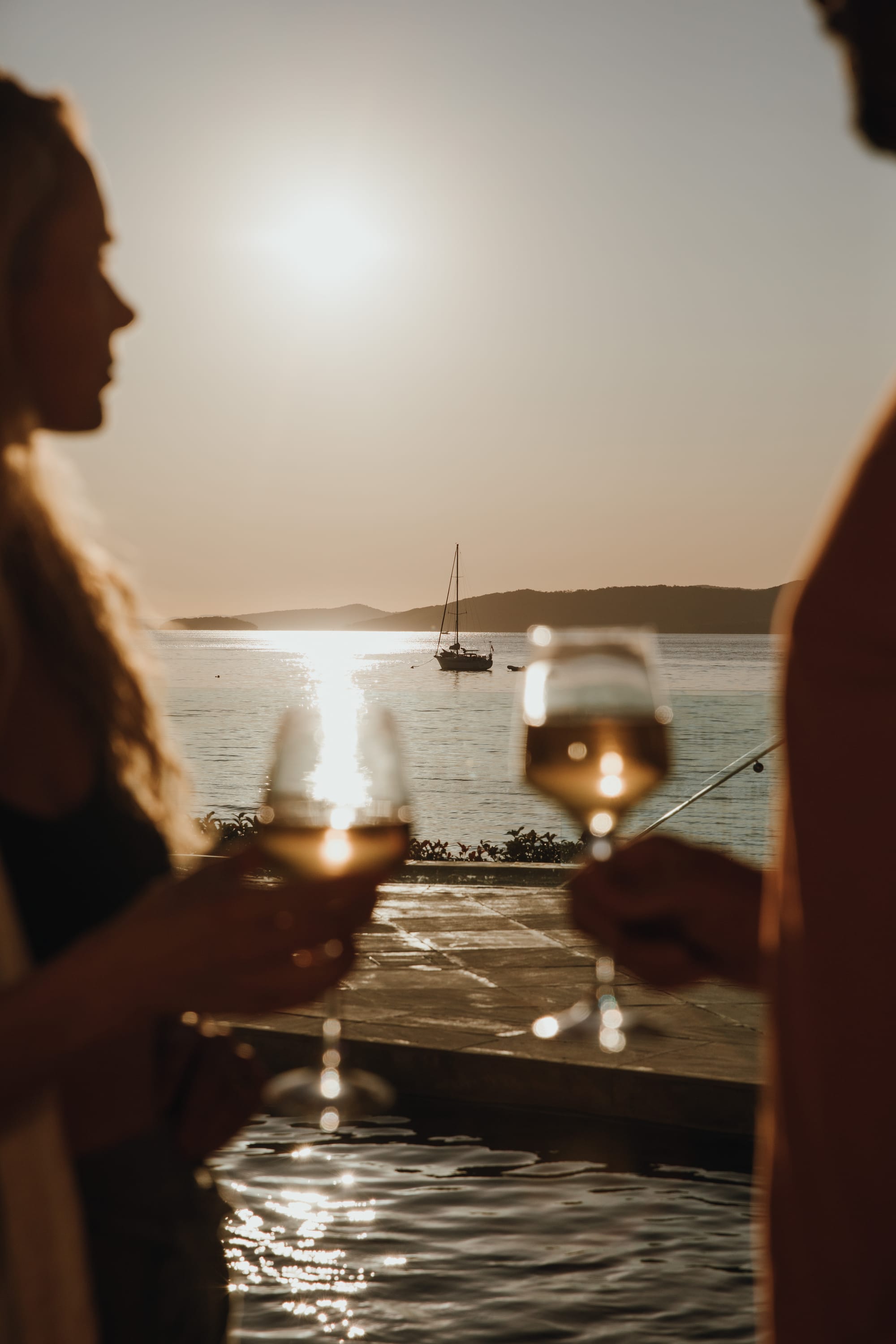 Amarna Luxury Beach Resort. Photography by Will Salkeld. Two people in foreground with two wine glasses. Background shows a sailboat on open water. 