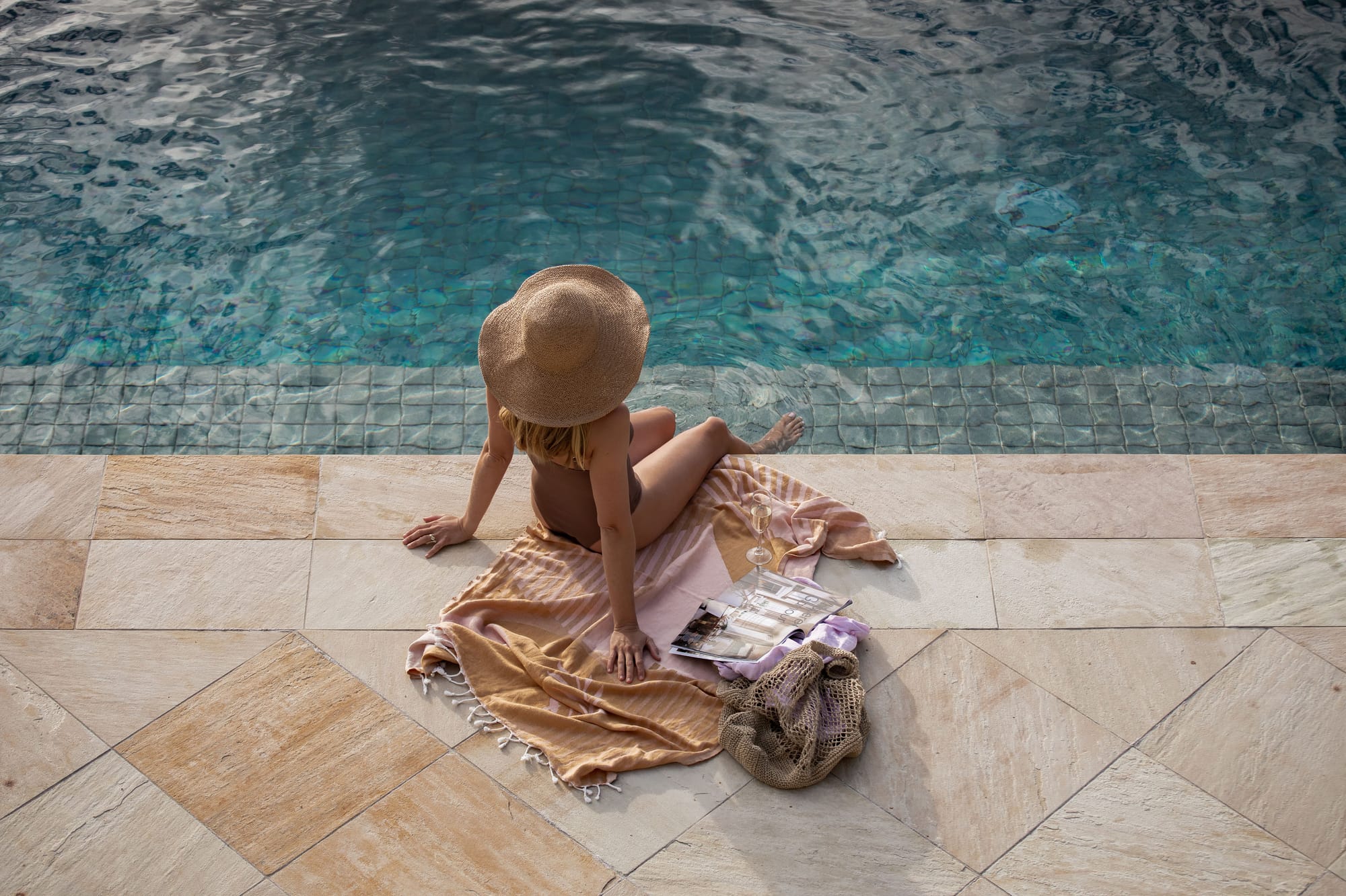 Amarna Luxury Beach Resort. Photography by Will Salkeld. Birds eye view of woman sitting on a towel with a large hat, with her feet in a tiled pool. Beige floor tiles outside of pool. 