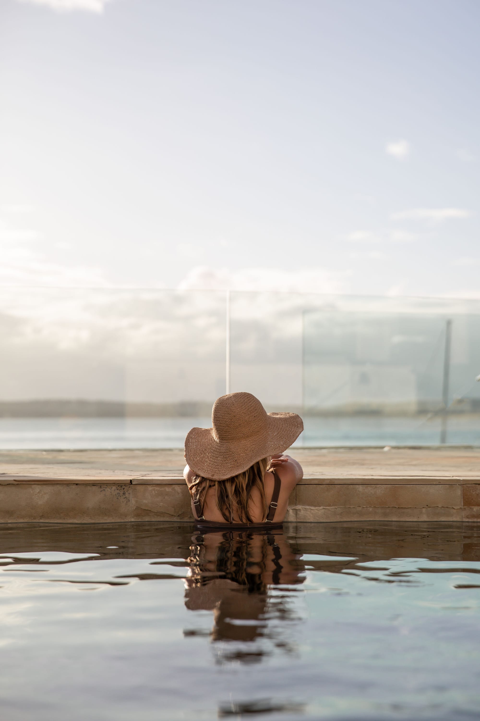 Amarna Luxury Beach Resort. Photography by Will Salkeld. Woman in sunhat leaning on stone edge of pool, in front of glass fence overlooking water.