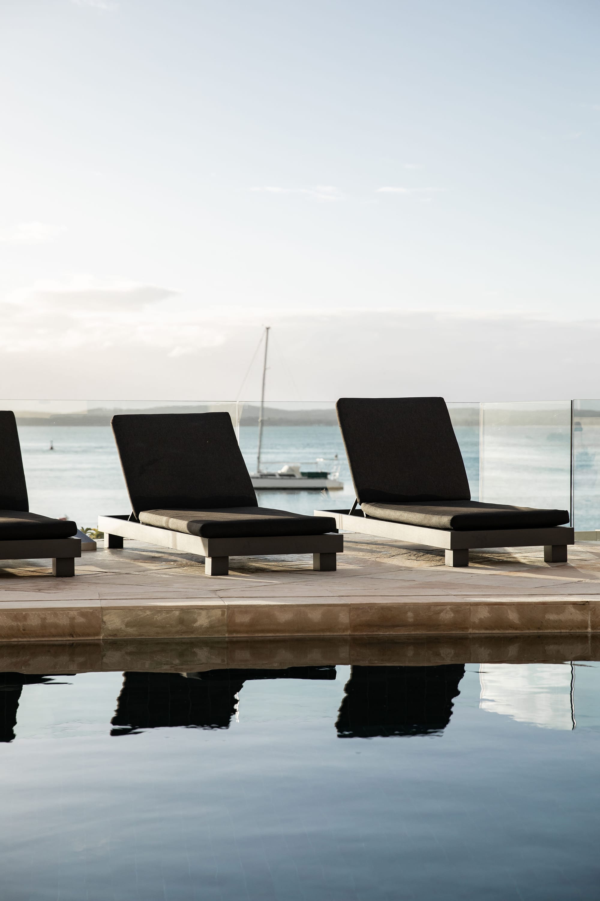 Amarna Luxury Beach Resort. Photography by Will Salkeld. Three black chaise lounges by pool, on beige floor tiles and in front of glass fence, which separates pool area from open harbor in background. 