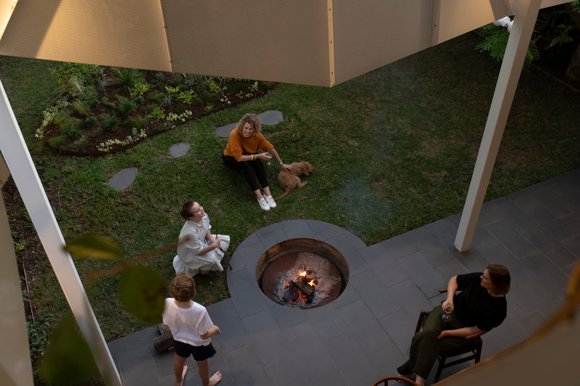 Cornerstone by Loupe Architecture. Photography by Alanna Jayne McTiernan. Birds eye view of family sitting around sunken firepit in outdoor courtyard, bordered by green grass. 