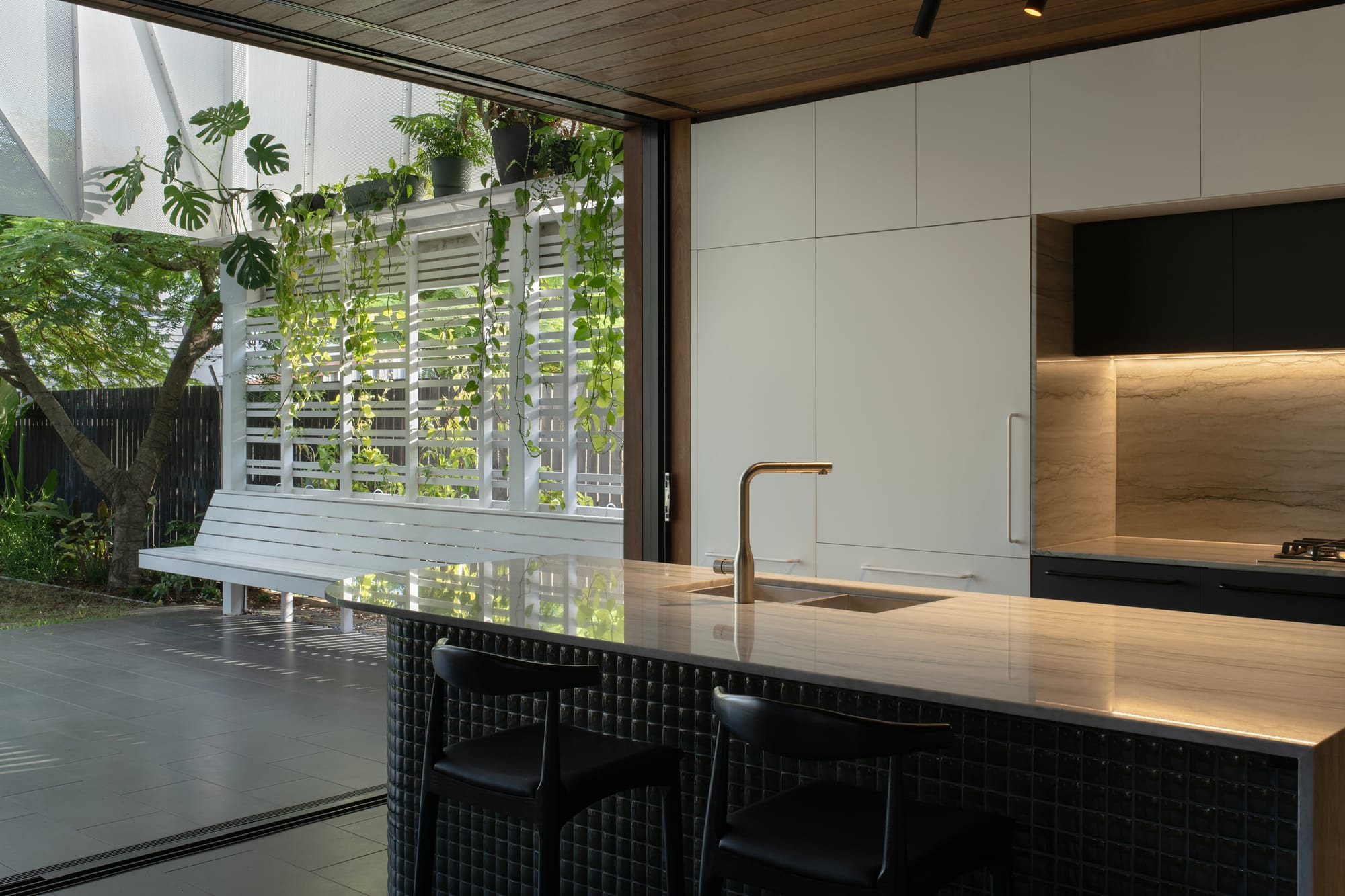 Cornerstone by Loupe Architecture. Photography by Alanna Jayne McTiernan. Kitchen overlooking outdoor patio with integrated white timber bench seat and screen with plants growing up it. 