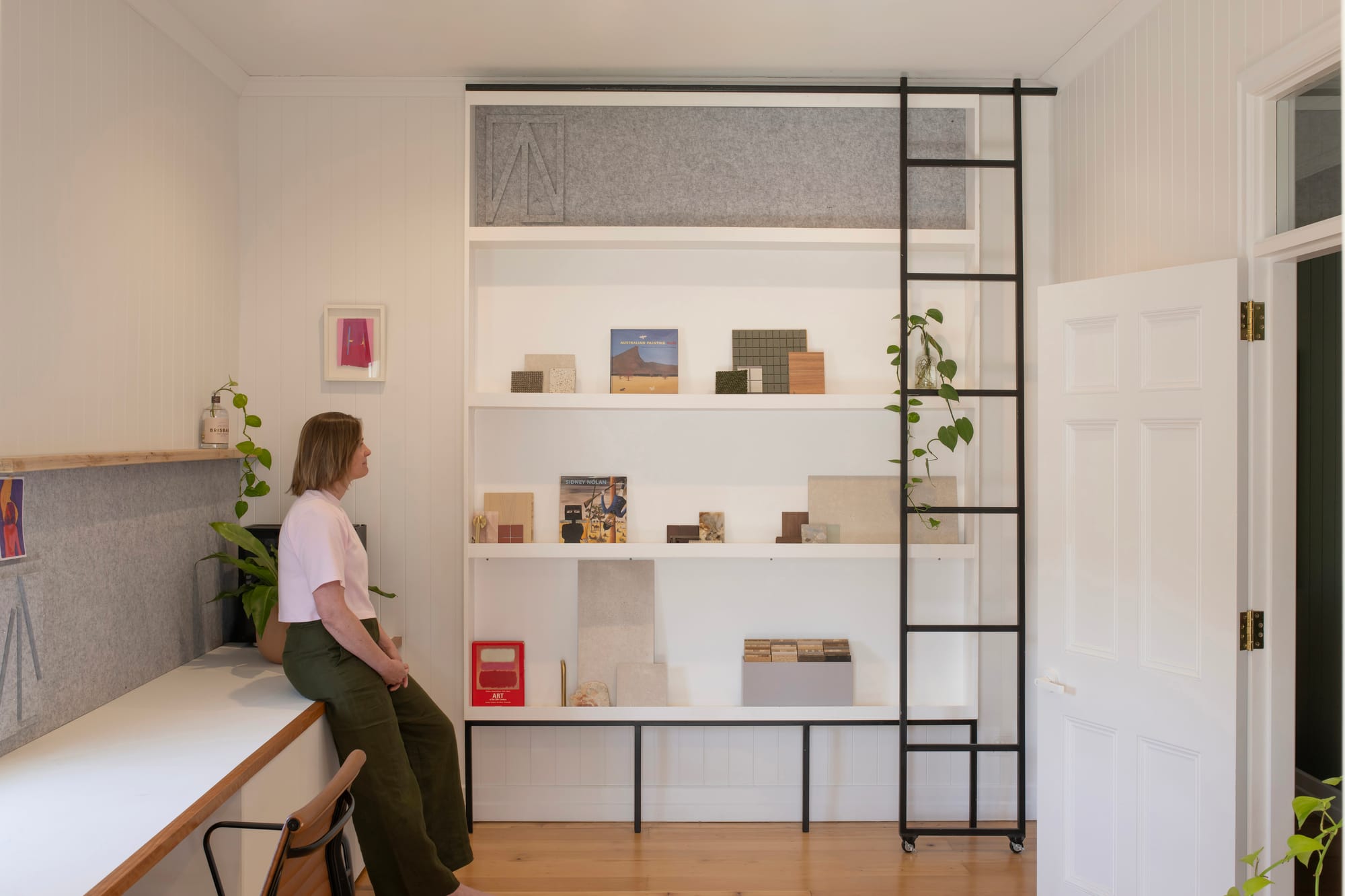 Cornerstone by Loupe Architecture. Photography by Alanna Jayne McTiernan. Integrated library wall with white cabinetry and black steel rolling ladder. Bench runs along length of right wall. 