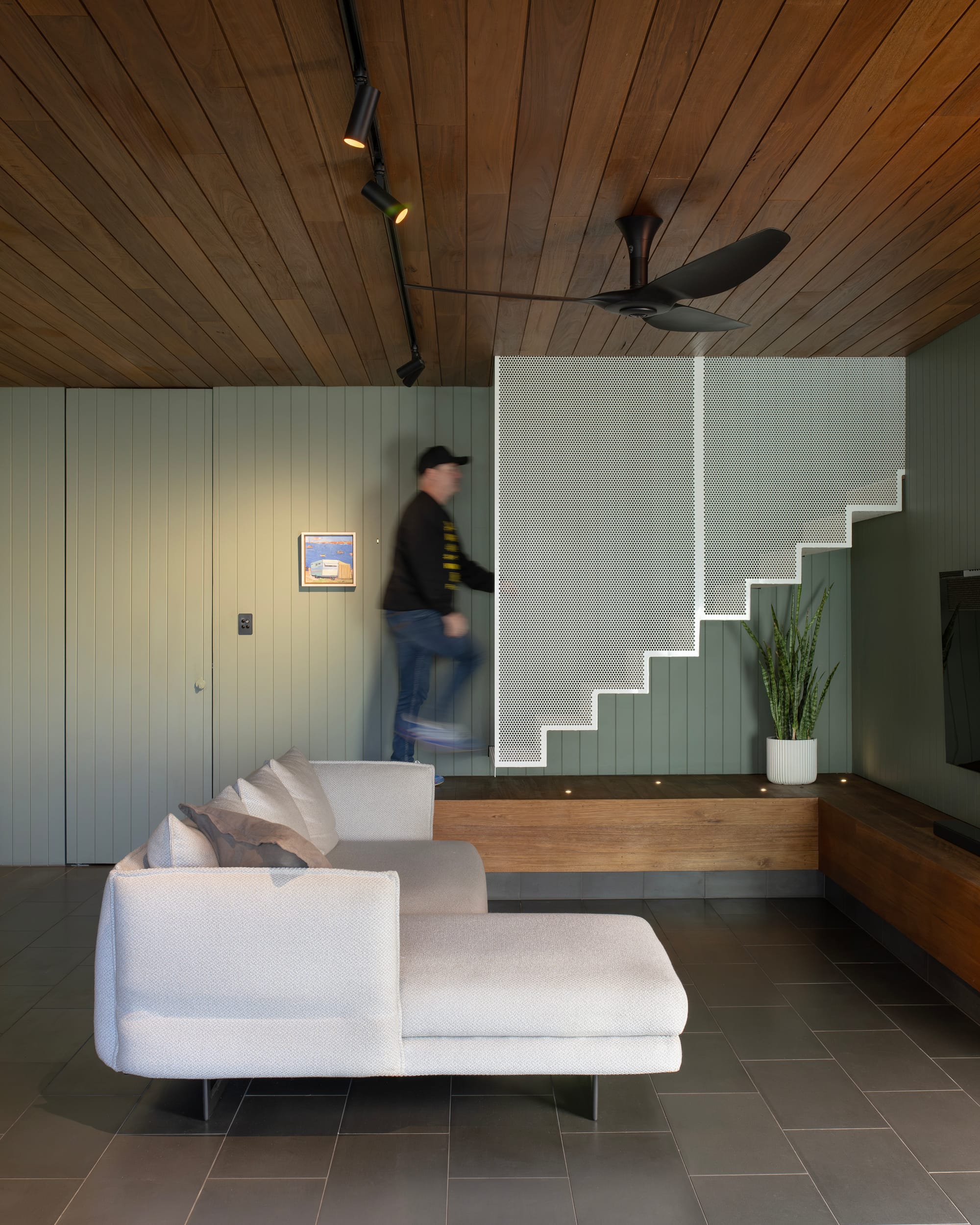 Cornerstone by Loupe Architecture. Photography by Alanna Jayne McTiernan. Living space with white modular lounge, bluestone tile floors, and a white perforated steel staircase and screen. Sage green timber clad walls. 