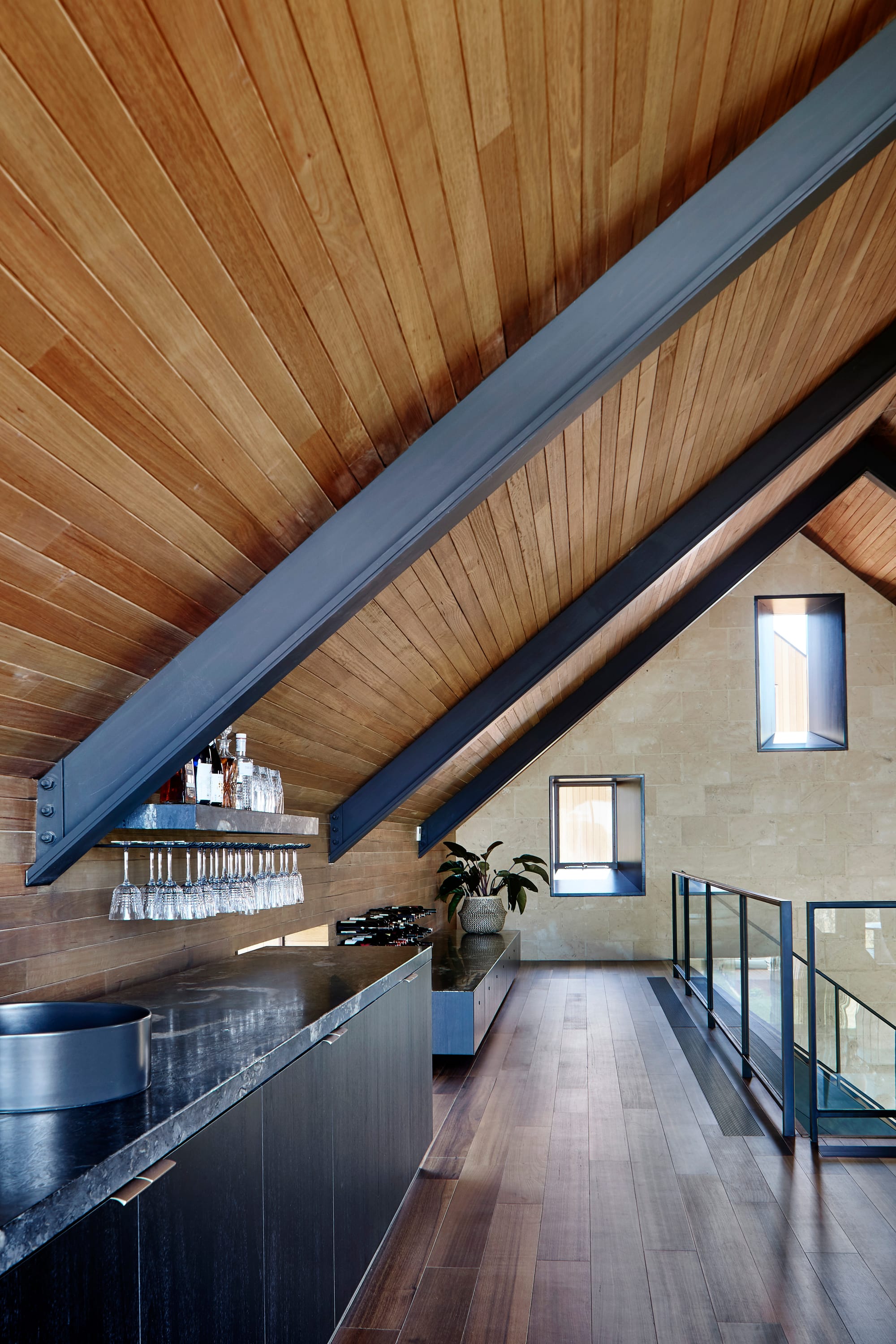 Tasmanian Timber in Flinders Residence by Abe McCarthy. Photography by Shannon McGrath. Loft with angled timber clad ceiling and timber floors. Bar kitchen with dark timber veneer runs along left pale brick wall.