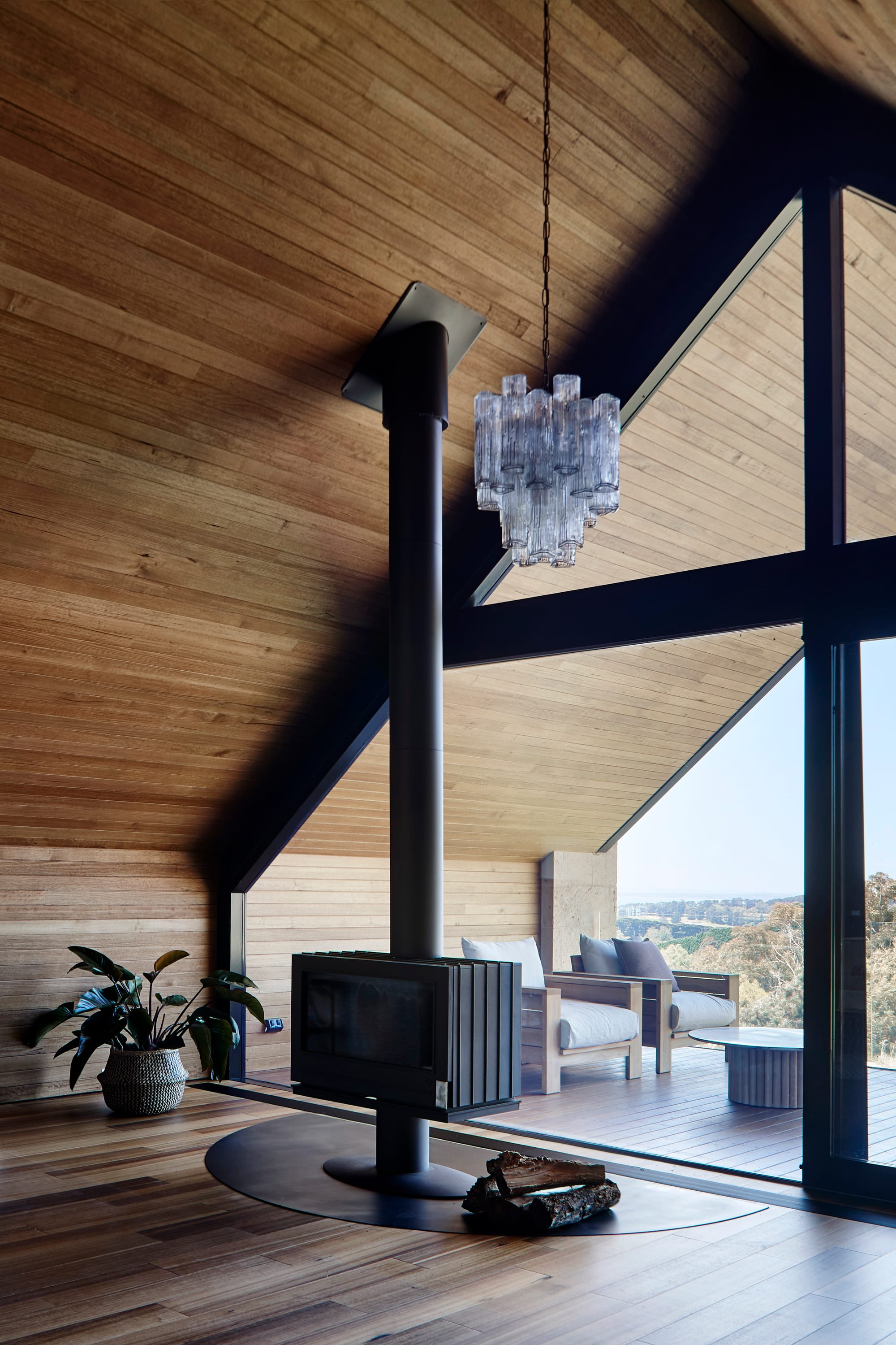 Tasmanian Timber in Flinders Residence by Abe McCarthy. Photography by Shannon McGrath. Floor-to-ceiling windows in tall, arched shape bordered by angled timber clad ceiling. Timber clad floors and black freestanding fireplace.