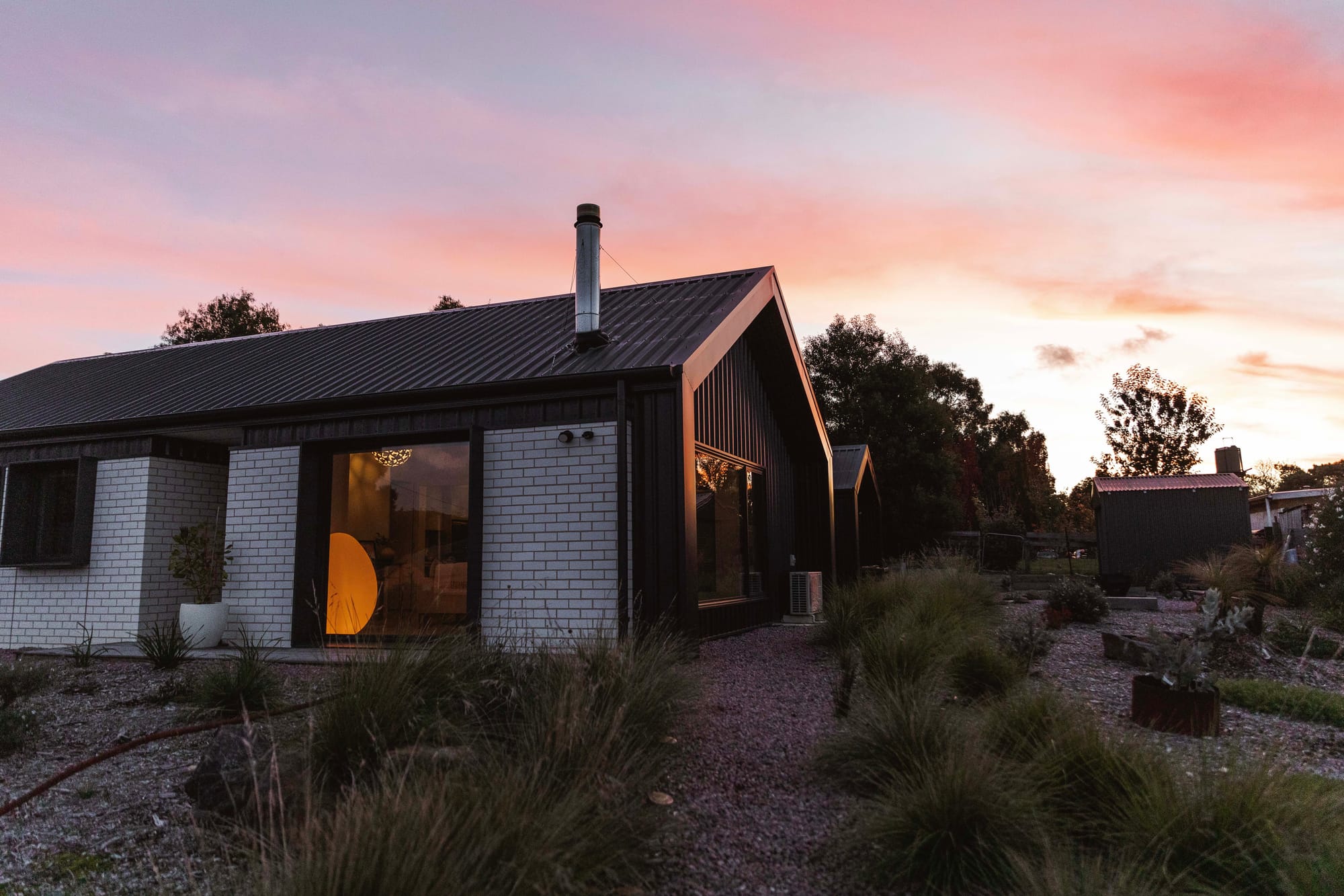 Forrest Haus Retreat. Images copyright of Forrest Haus Retreat. White brick home with black steel roof in front of pink sunset on a hill.