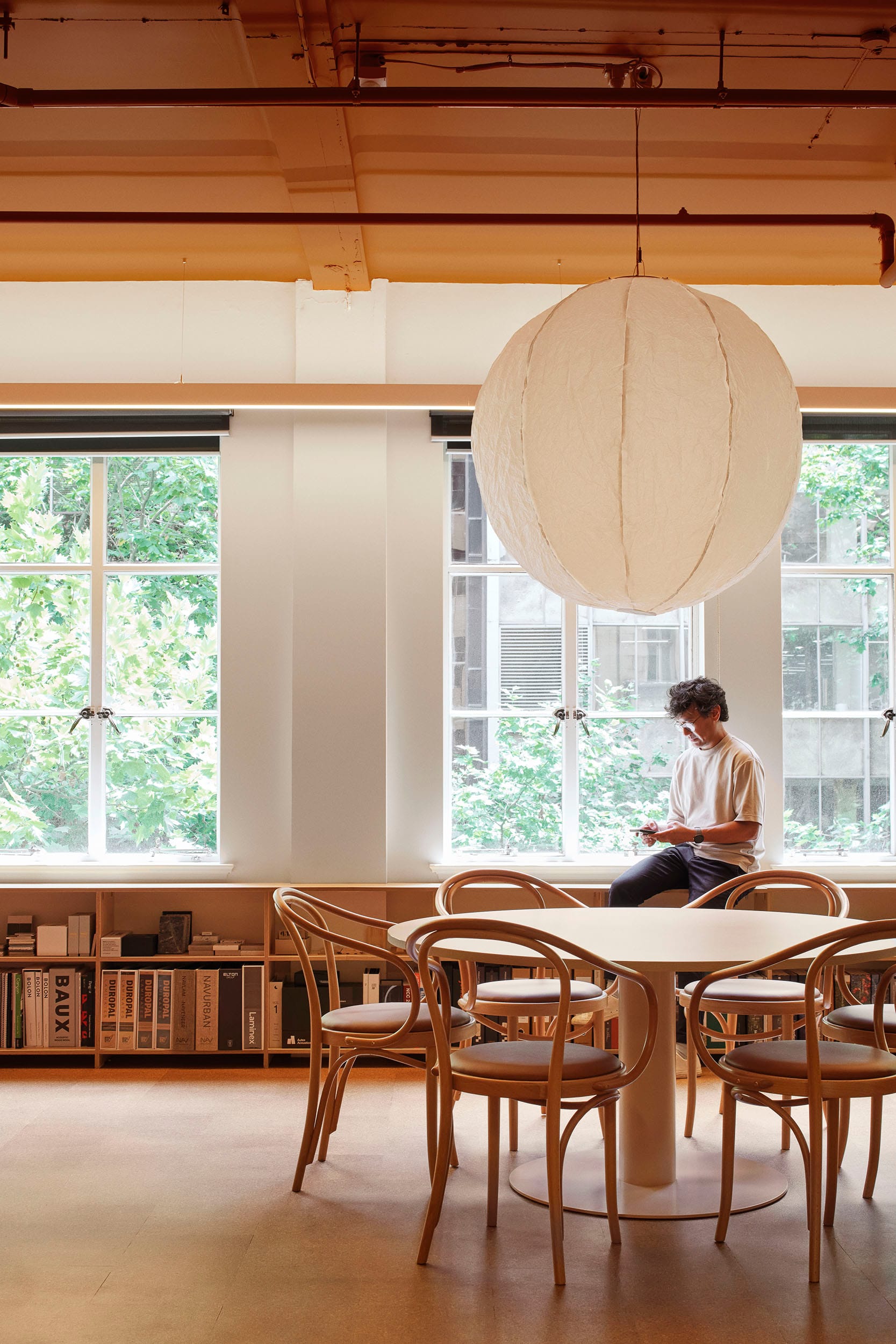 Hames Sharley Melbourne Studio by Hames Sharley. Photography by Nicole England. Round timber table in office staff room with large white pendant light. Low bookcases run along window.