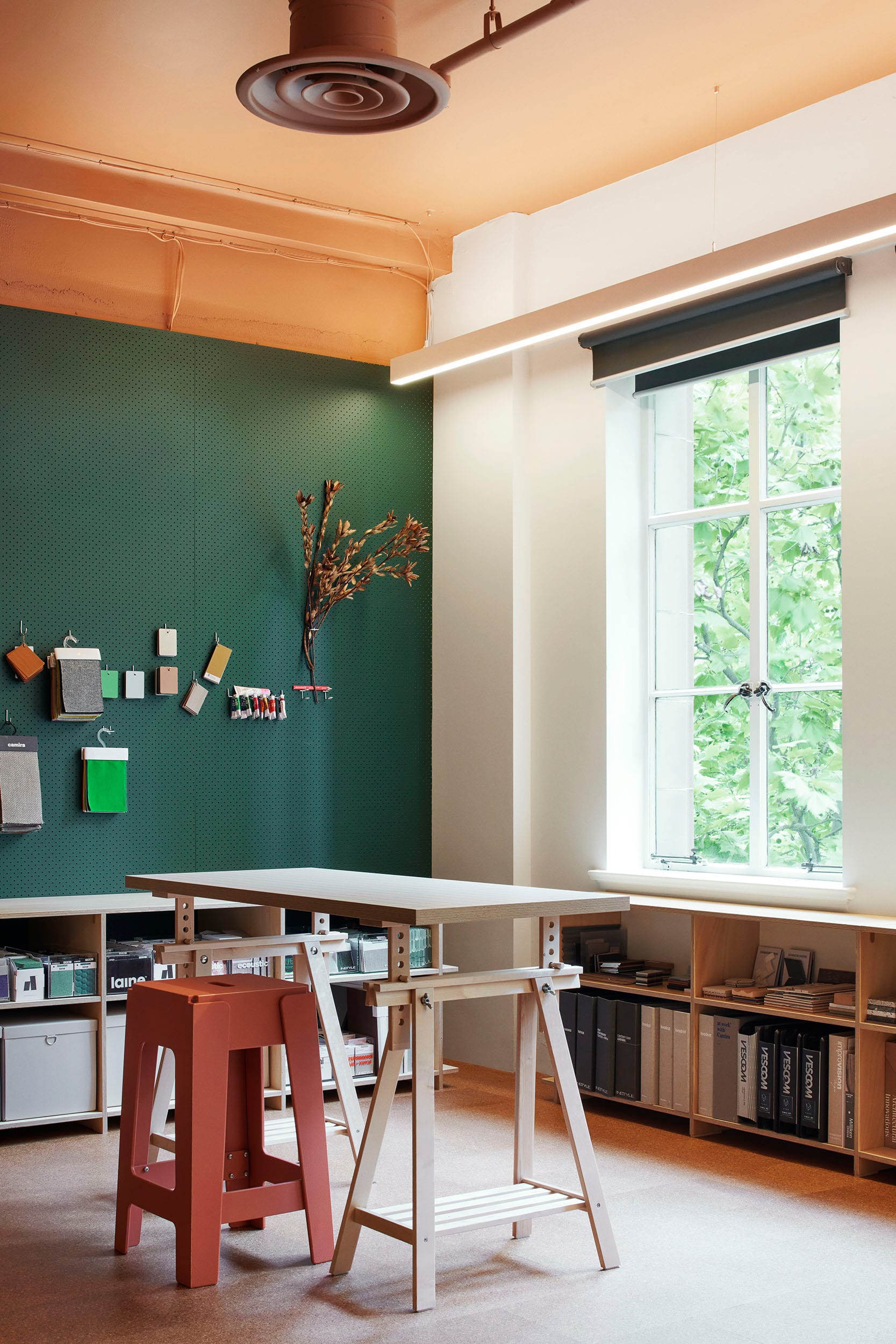 Hames Sharley Melbourne Studio by Hames Sharley. Photography by Nicole England. Makerspace with white trestle table and red stool. Dark green moss pin wall and cork floors.