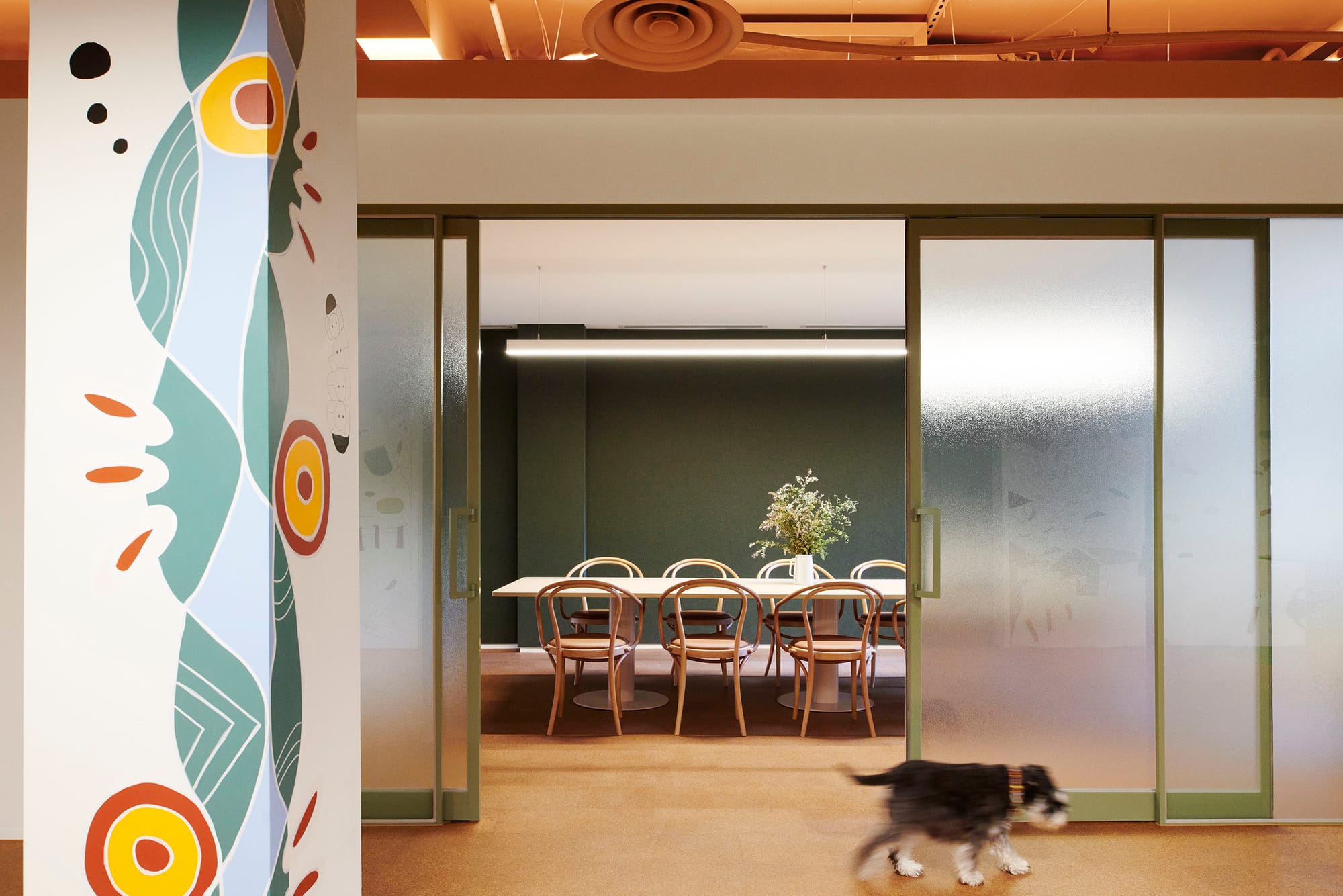 Hames Sharley Melbourne Studio by Hames Sharley. Photography by Nicole England. Meeting space in office with gren walls, green framed windowed doors and long timber table. Mural on pillar in front.