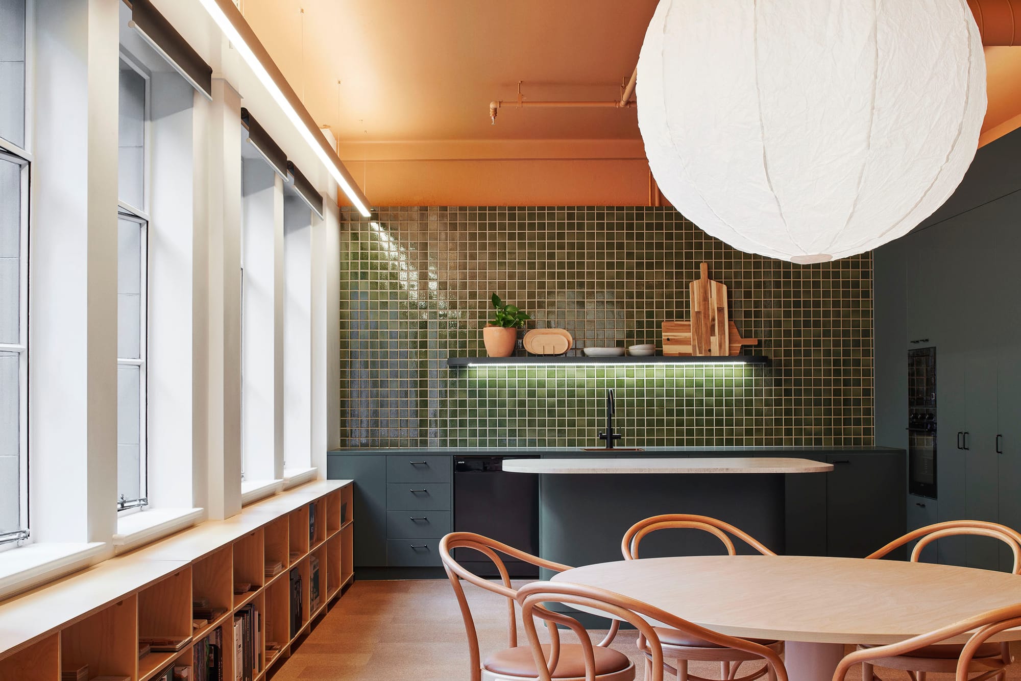 Hames Sharley Melbourne Studio by Hames Sharley. Photography by Nicole England. Office kitchen space with dark teal kitchen cabinetry and green mosaic tiles. Large white pendant light over round timber table. 