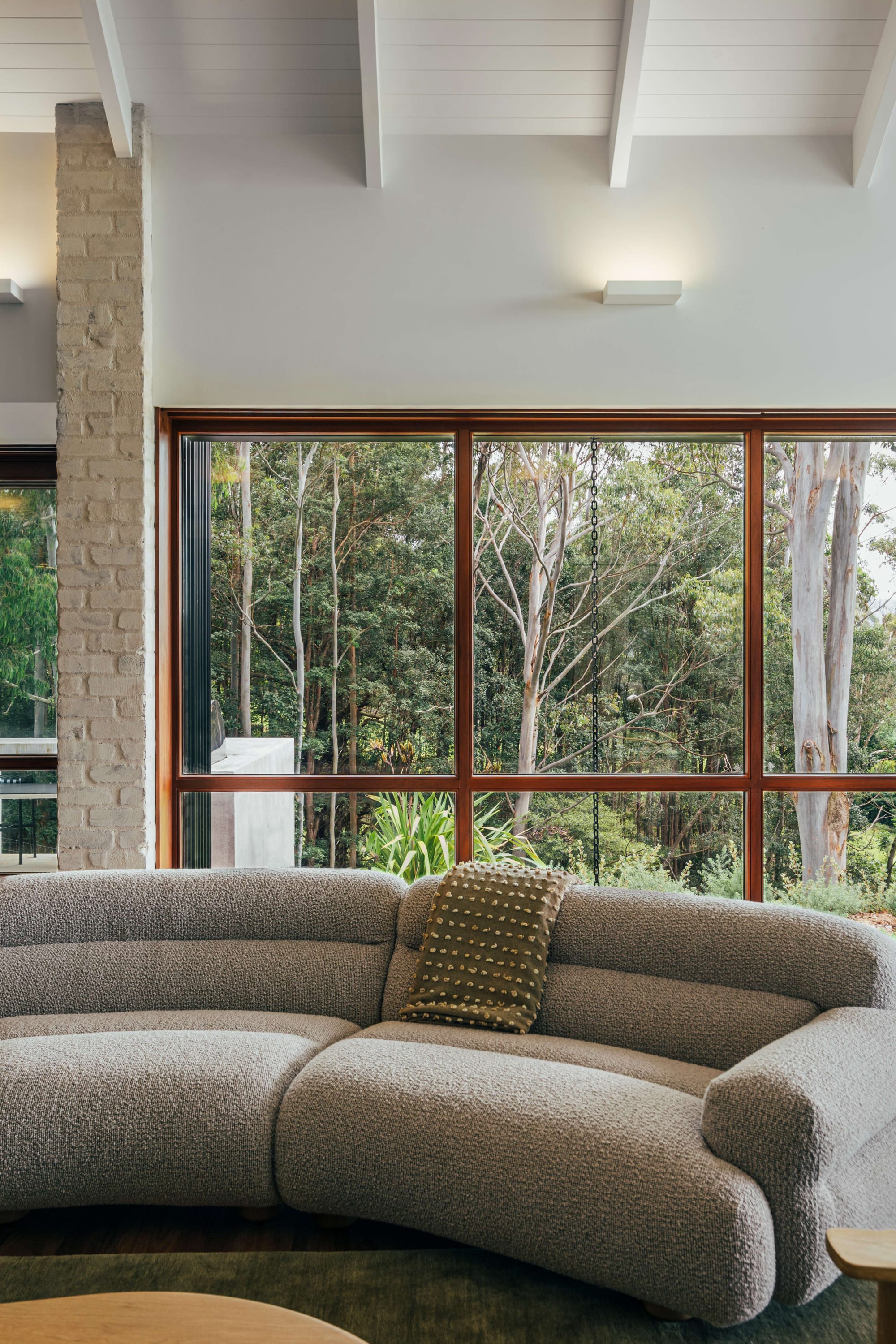 Project 465 by Robertson Collectif. Photography by Anna Mul. Living space with curved grey modular couch in front of full heigh windows overlooking bushland. 