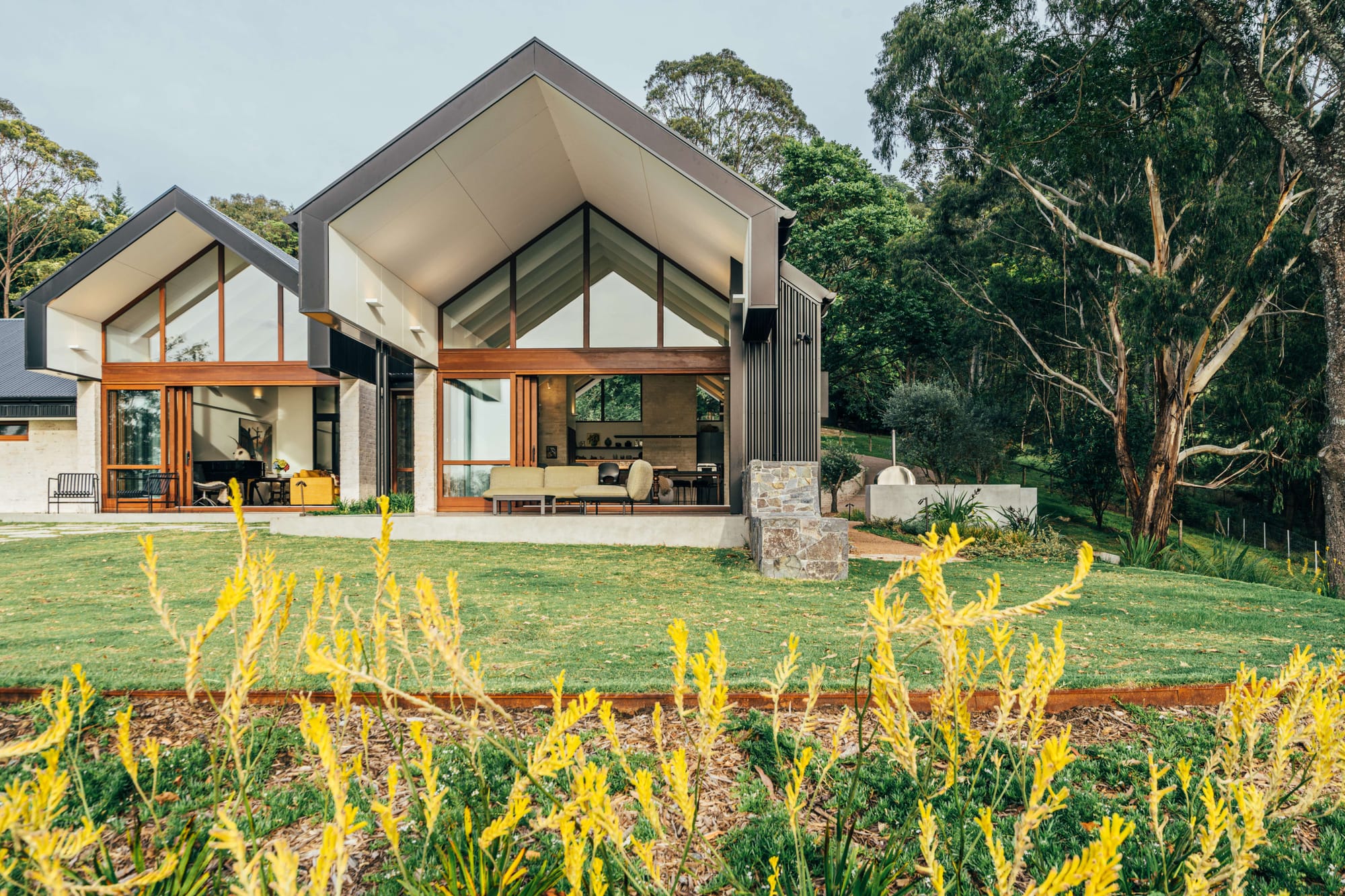 Project 465 by Robertson Collectif. Photography by Anna Mul. Rear facade of home with tall gable roofs, multiple patios and sprawling green garden. Native Australian bush in right hand background. 