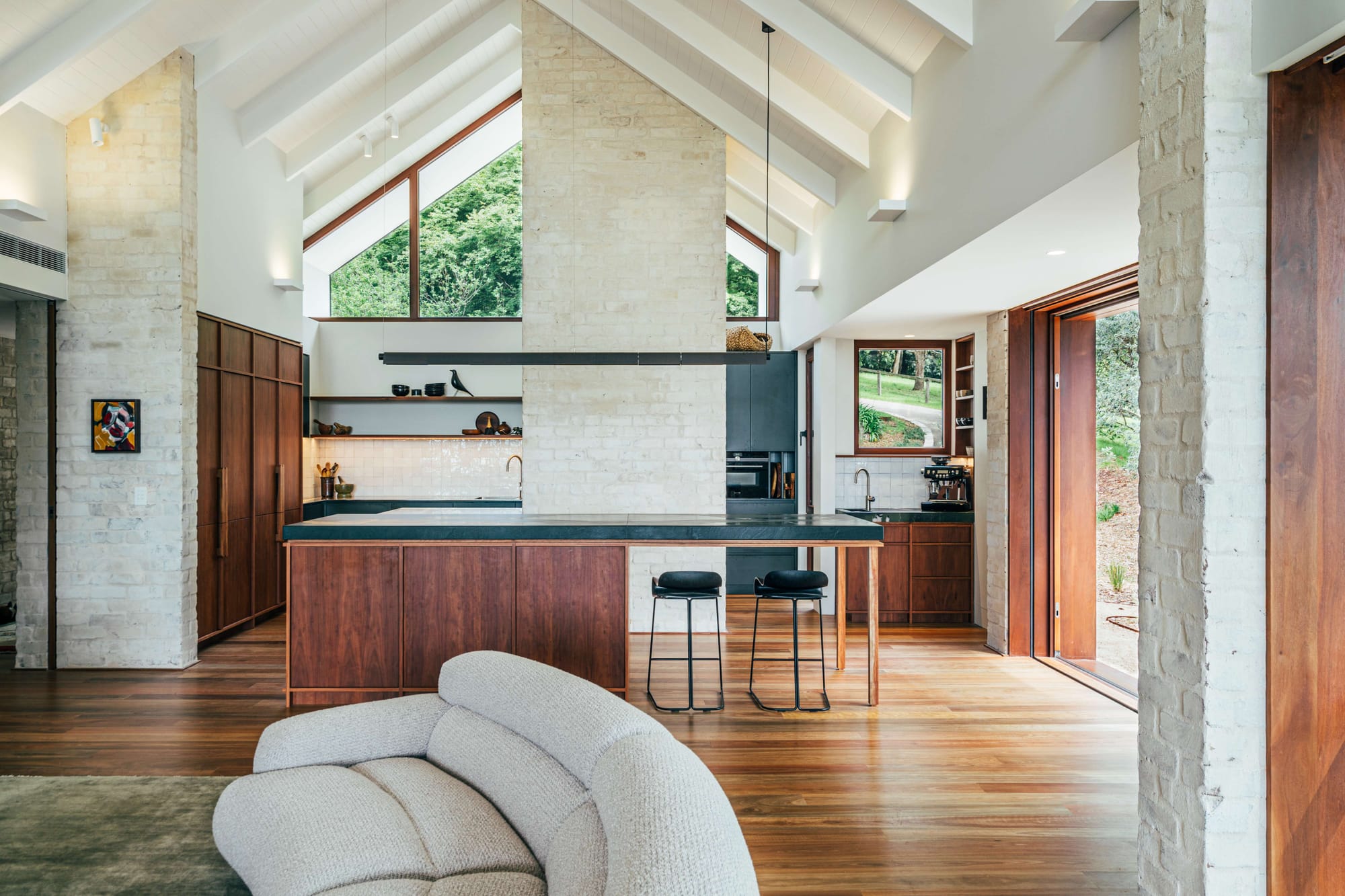 Project 465 by Robertson Collectif. Photography by Anna Mul. Open plan kitchen and living space with timber floors and gabled ceiling. White brick walls and curved grey couch. 