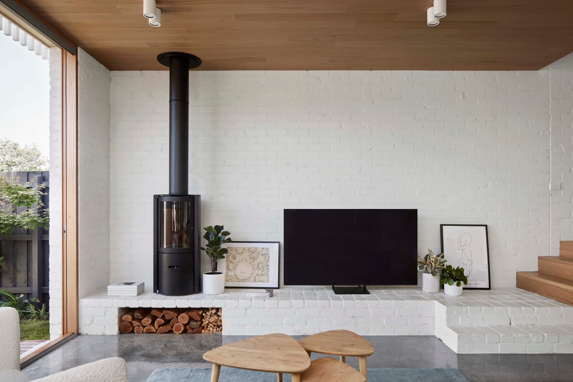 Rosie by Eckersley Architects. Photography by Tess Kelly. Living space with brick podium with freestanding black fireplace. Polished concrete floor, timber ceilings and white brick walls. 