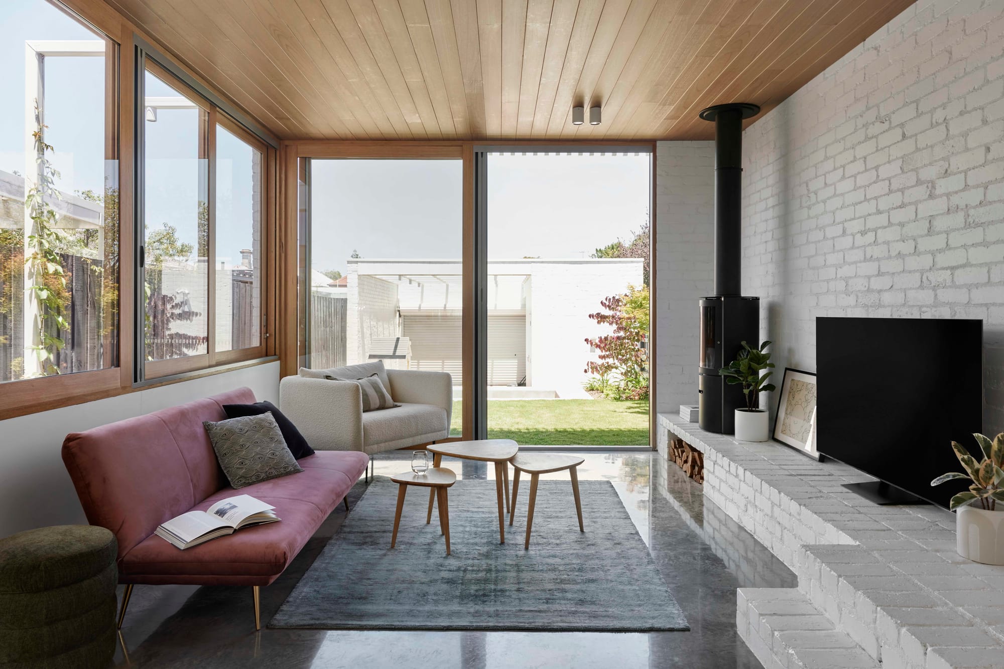 Rosie by Eckersley Architects. Photography by Tess Kelly. Living space with white brick walls, timber ceiling and polished concrete floor. Purple couch and sliding timber frame door leading to grassy backyard. 