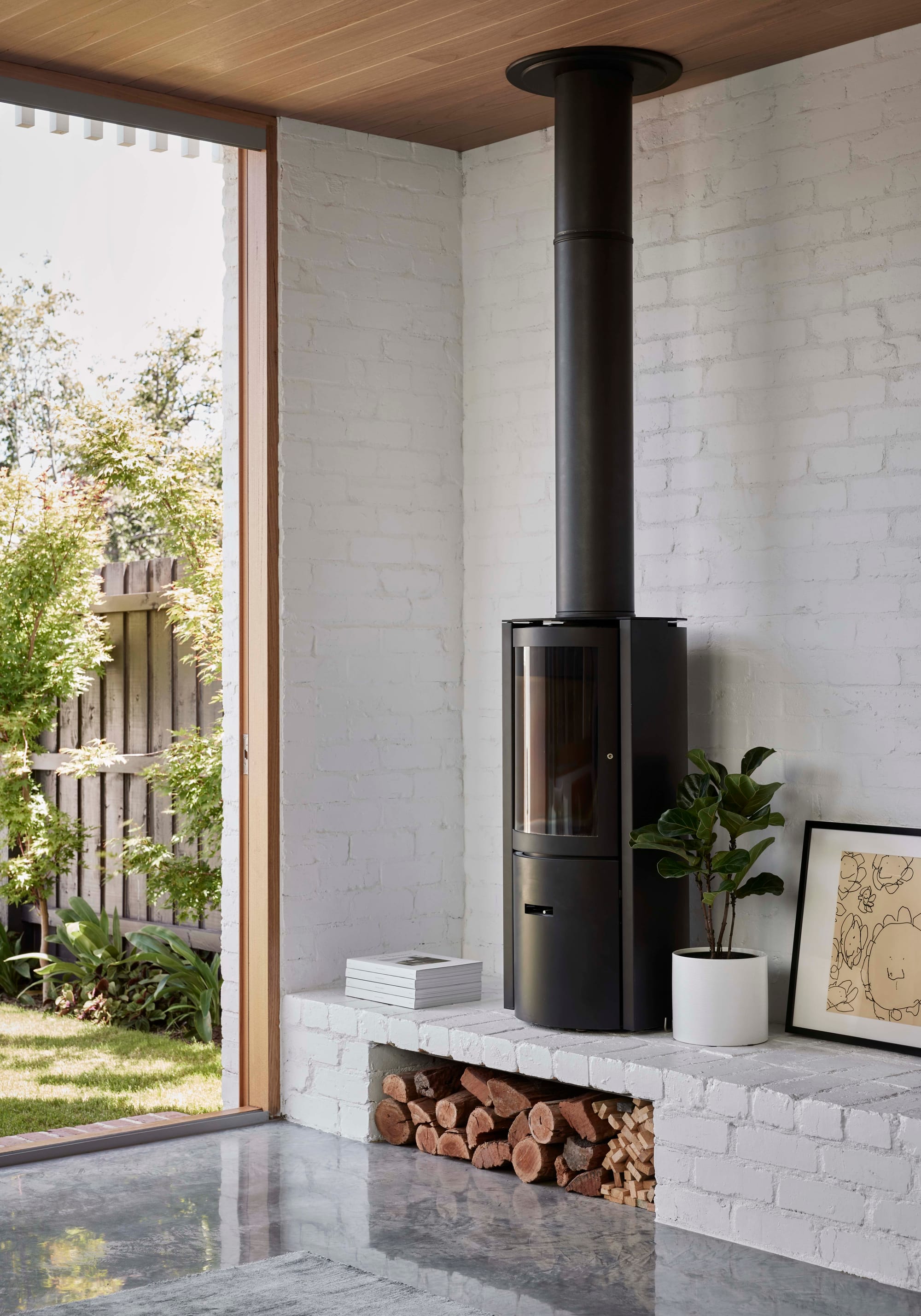 Rosie by Eckersley Architects. Photography by Tess Kelly. White brick wall interior with polished concrete floor and black freestanding fireplace. Timber framed door opens onto backyard. 