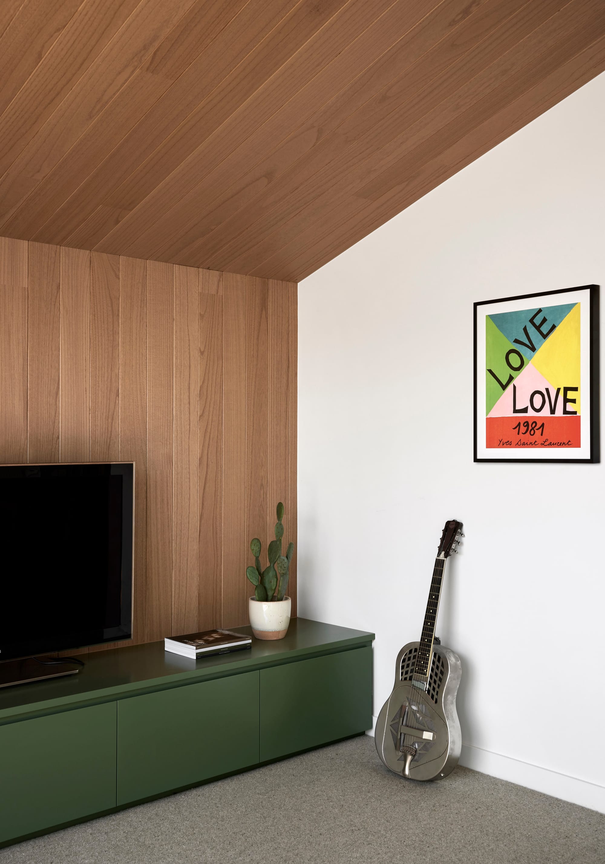 Rosie by Eckersley Architects. Photography by Tess Kelly. Corner of living space with timber walls and gabled ceiling, grey carpet and green integrated storage. Silver guitar leaning on white wall. 