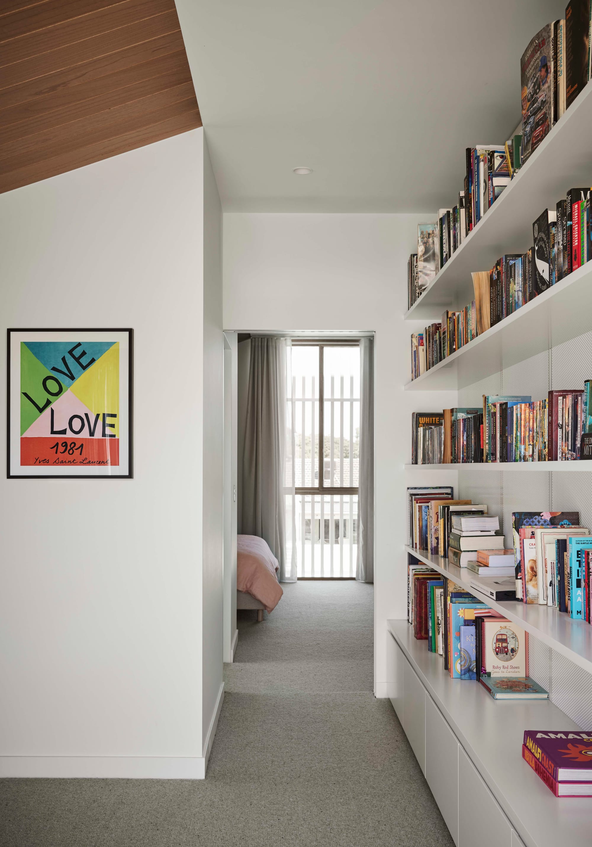 Rosie by Eckersley Architects. Photography by Tess Kelly.  Integrated white bookshelf running along hallway door, leading to bedroom with grey carpets.