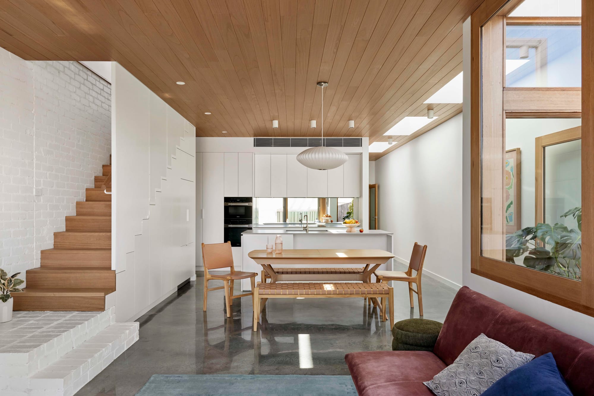 Rosie by Eckersley Architects. Photography by Tess Kelly. Open plan living space with polished concrete floors, timber ceiling and white walls. Timber staircase, windows frames and dining table. 