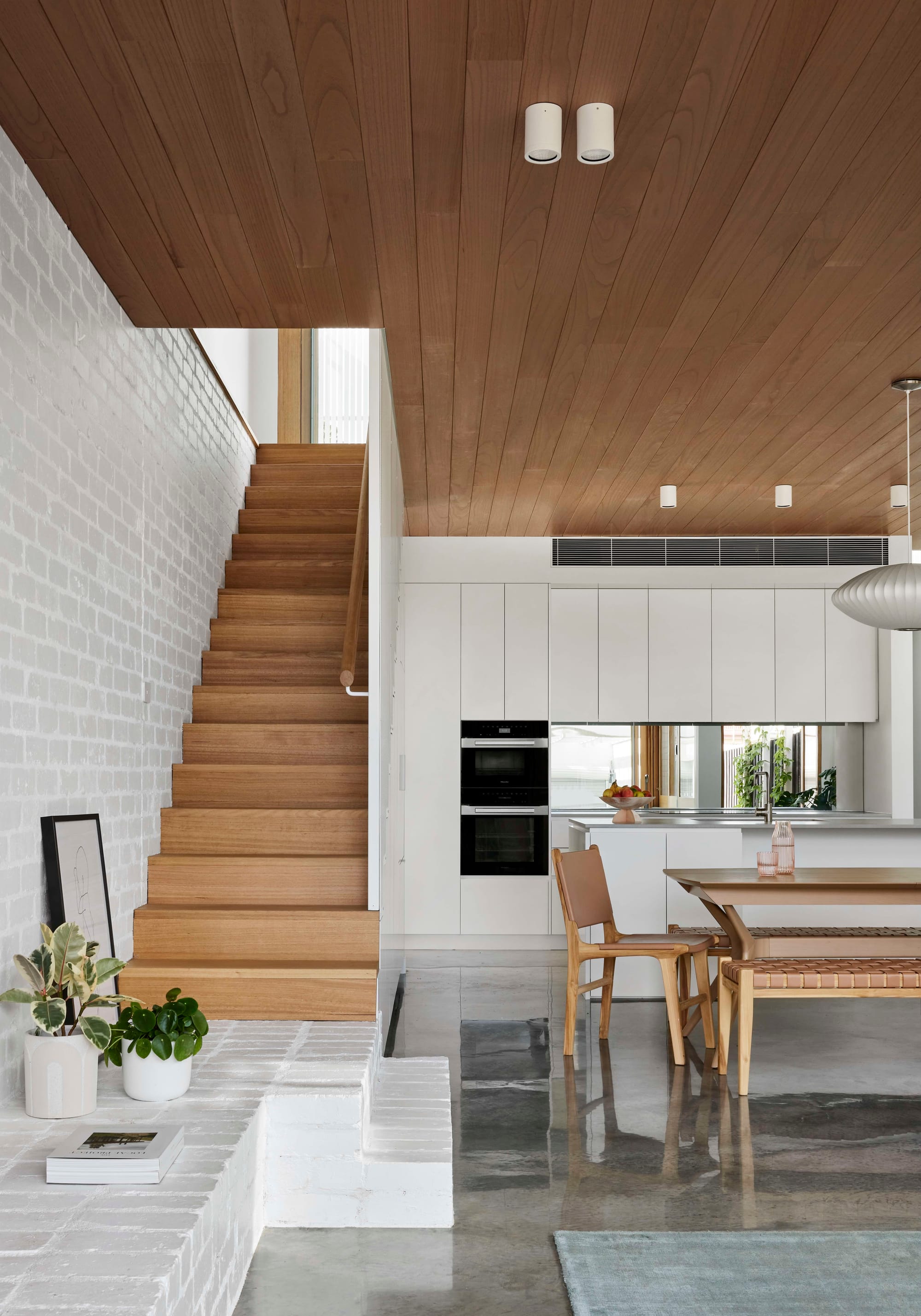 Rosie by Eckersley Architects. Photography by Tess Kelly. Timber staircase leading away from ground floor open plan space with polished concrete floors and white brick walls. 