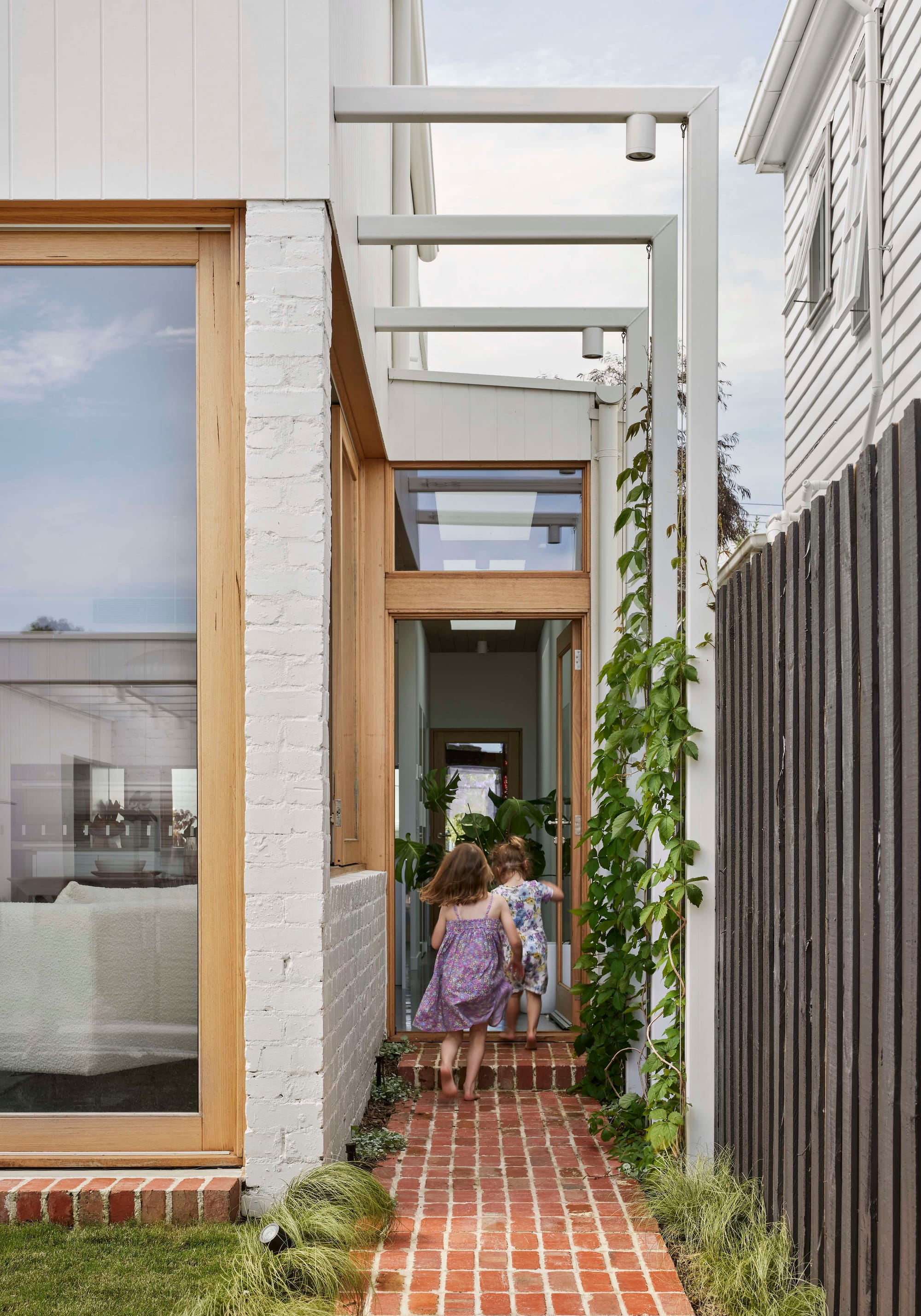 Rosie by Eckersley Architects. Photography by Tess Kelly. Children running up brick footpath to timber framed door leading into residential home. Green crawling plants grow on left fence.