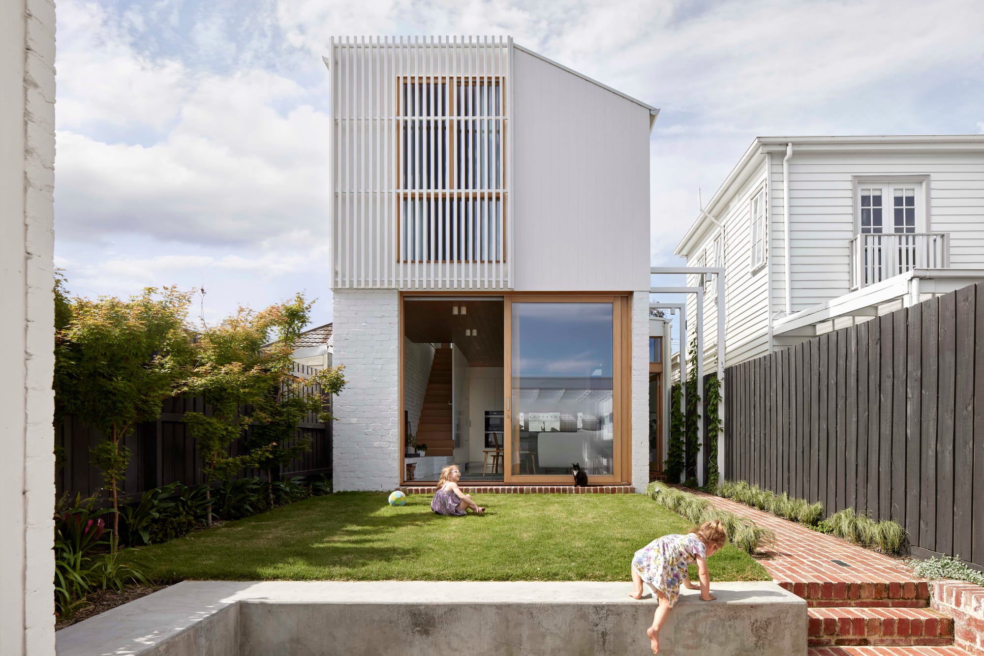 Rosie by Eckersley Architects. Photography by Tess Kelly. Rear facade of double storey white home with green grass backyard and concrete retaining wall.