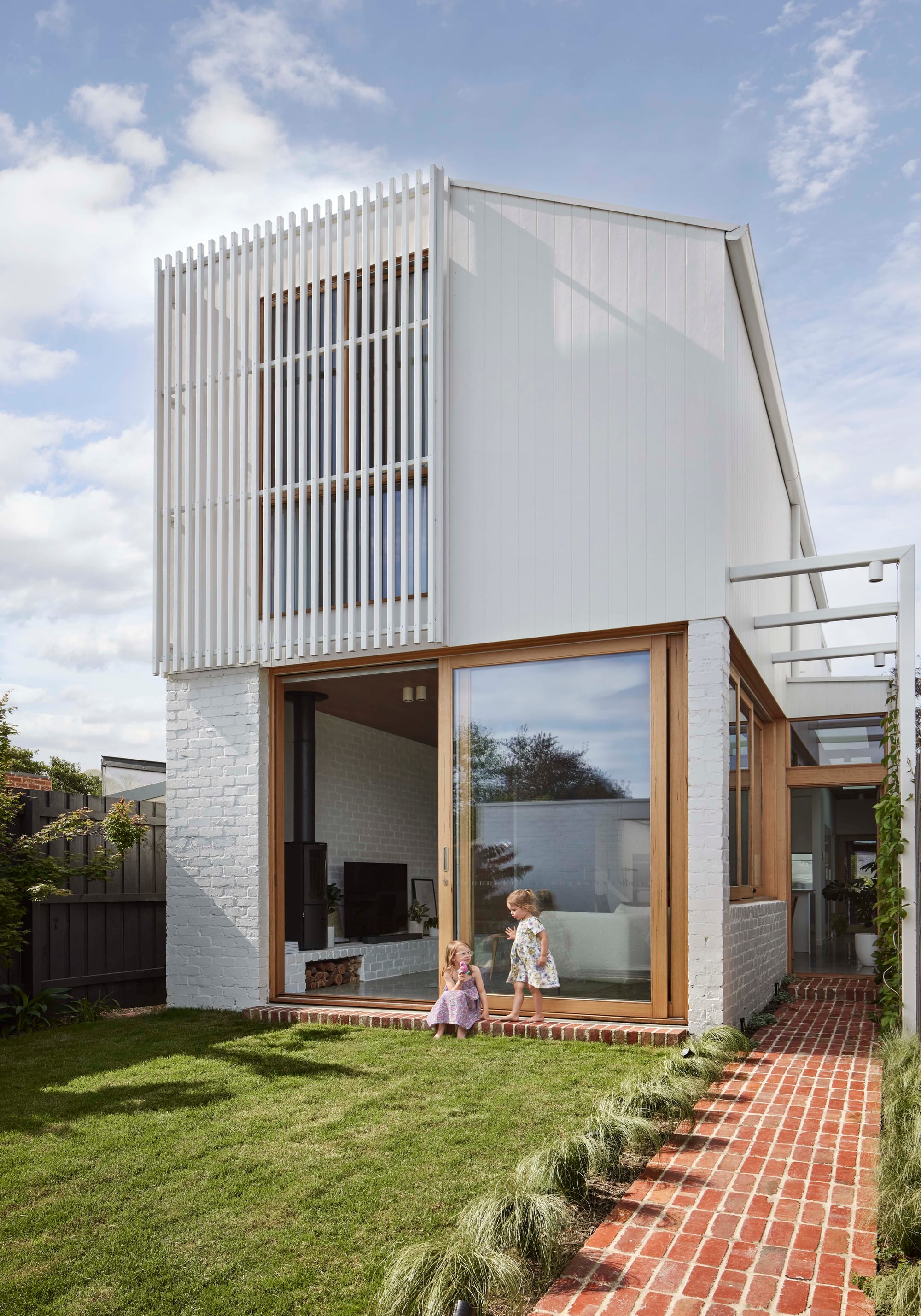 Rosie by Eckersley Architects. Photography by Tess Kelly. Rear facade of home with brick pathway leading to timber framed doorway. Green grass backyard and white brick walls.