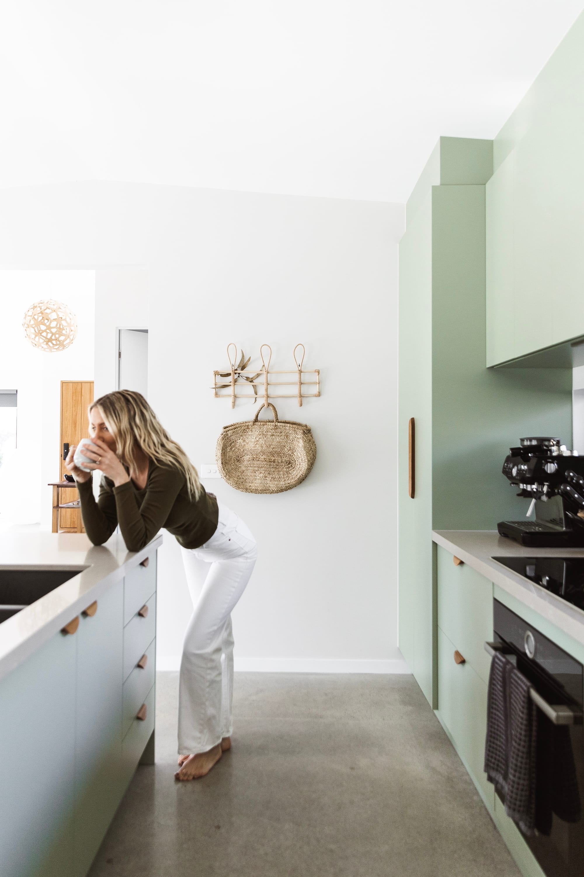 Forrest Haus Retreat. Images copyright of Forrest Haus Retreat. Light mint kitchen with high white ceilings and two geometric pendants. An interior shot of the kitchen showing a woman leaning over the bench top and drinking coffee