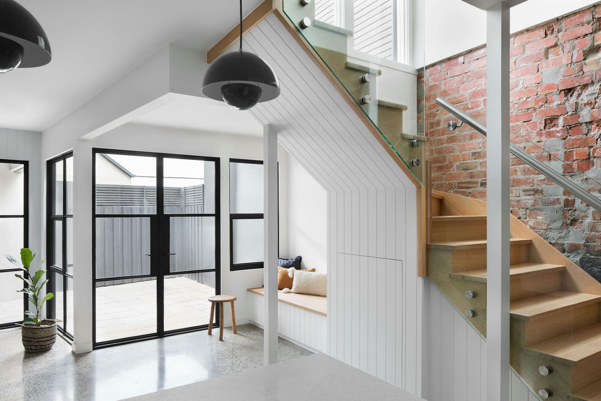 Stead House by Hannaford Design Studio. Entrance area with outside view, featuring metal frame glass door, wooden staircase with brick wall.