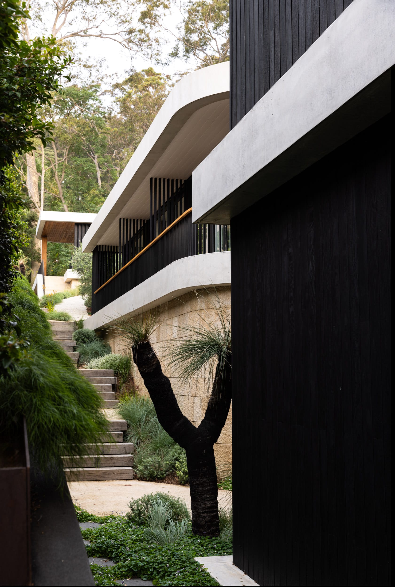 Ripple House by Marker Architecture. Exterior view on house with stained black wood panels and sandstone walls surrounded by nature.