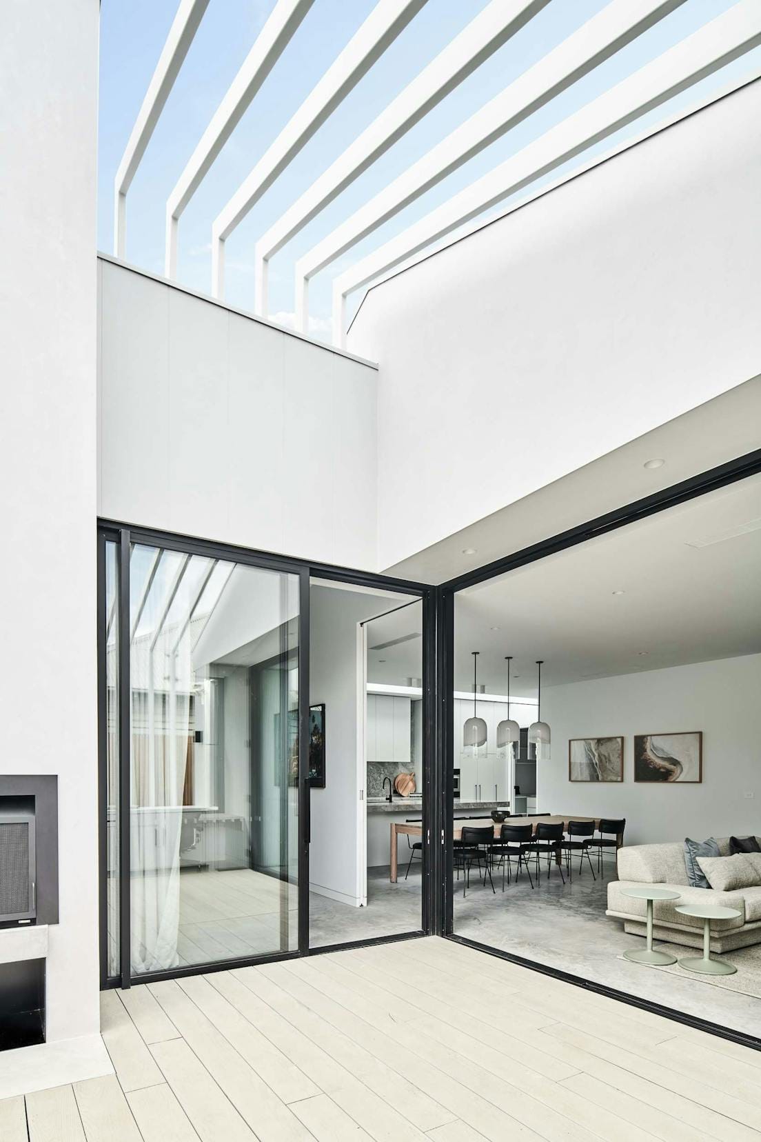 Elsterwick House by Merrylees Architects. View to interior featuring beige sofa and kitchen area with wooden table and black dining chairs.