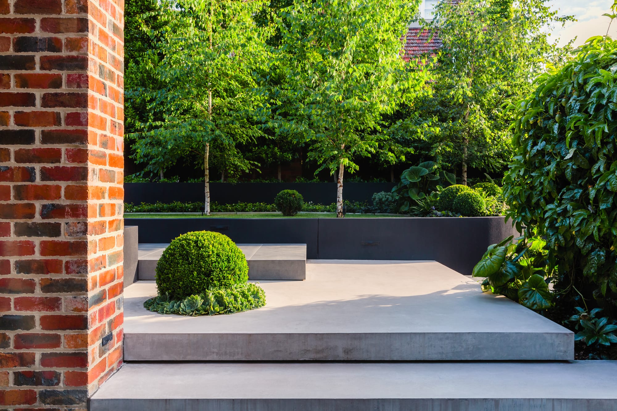 Garden featuring layered concrete hardscaping and stone staircase and red brick walls.