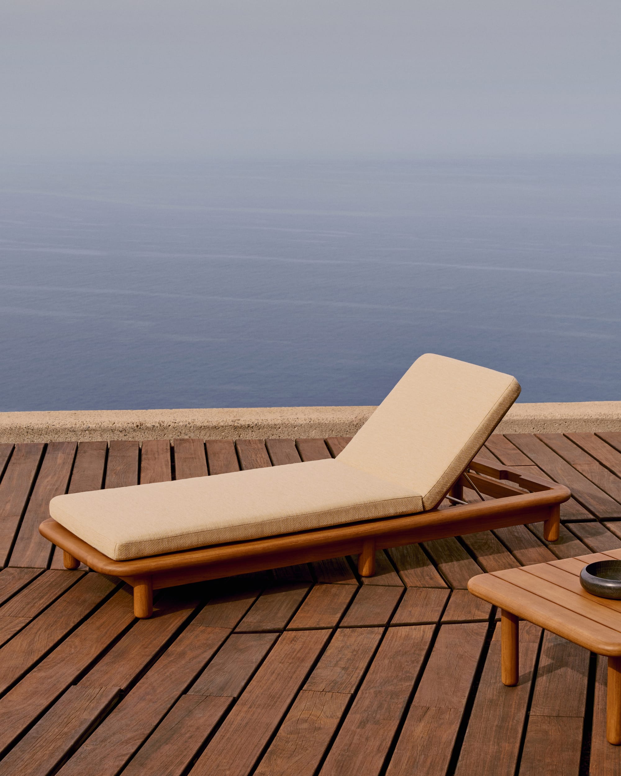 Turquetta by Kave Home. Curved teak wood outdoor sun bed featuring beige accent fabric, coffee table and ocean view.