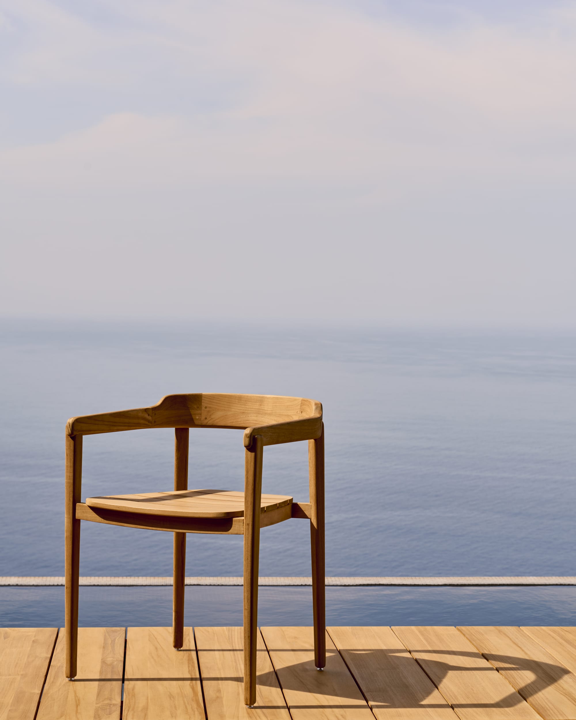 Icaro by Kave Home. Modern teak wood outdoor chair on Mediterranean terrace with view on infinity pool and ocean.