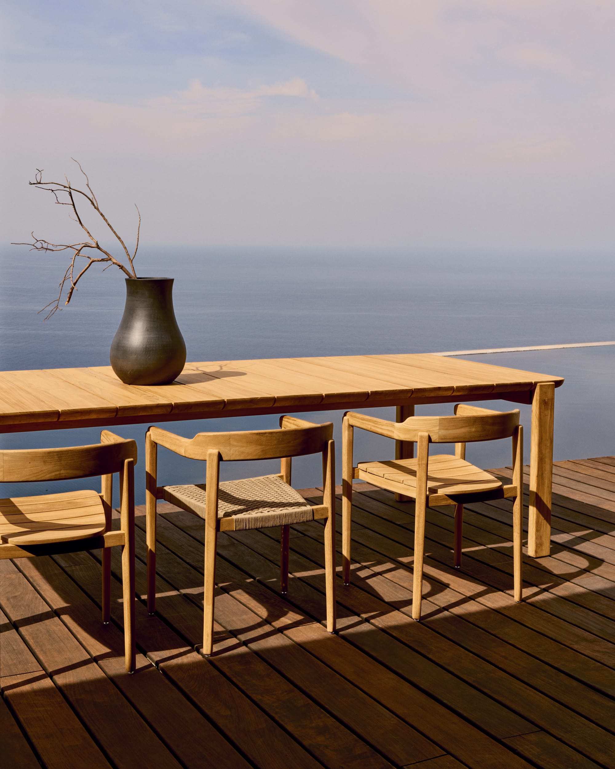 Icaro by Kave Home. Modern teak wood outdoor dining table set on Mediterranean terrace with view on infinity pool and ocean.