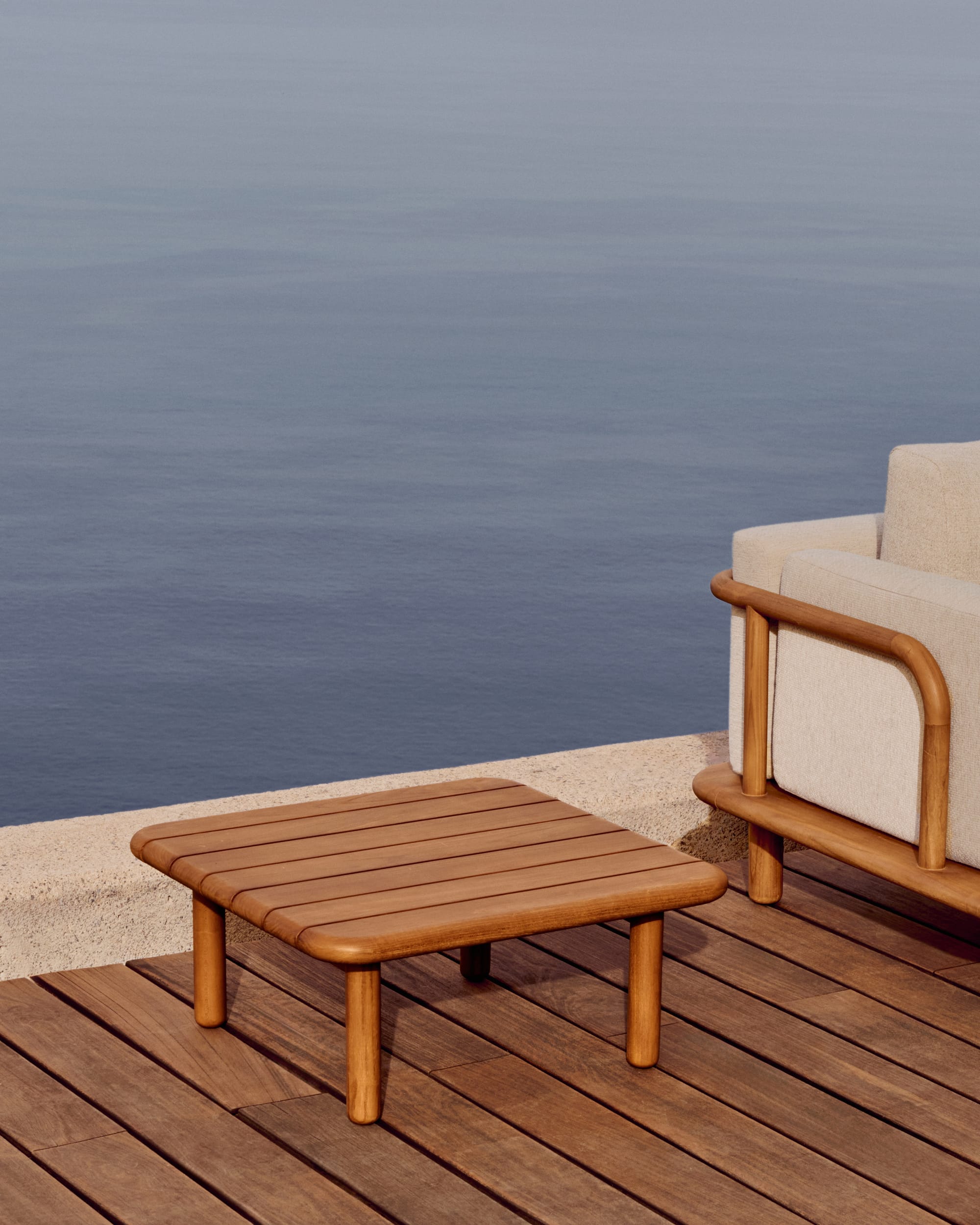 Turquetta by Kave Home. Curved teak wood outdoor coffee table and sofa featuring beige accent fabric and ocean view.