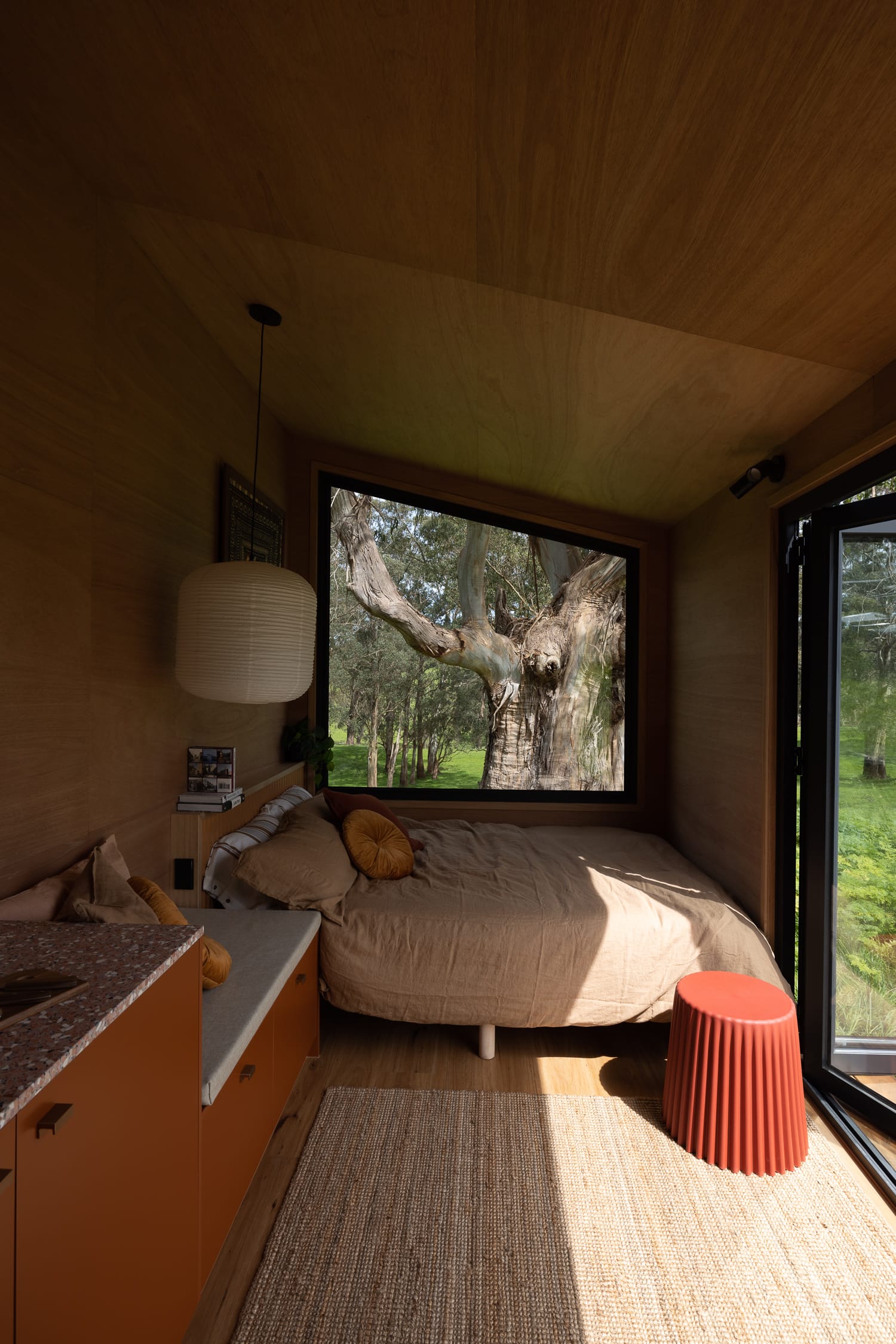 Shelby Lenehan Series by Base Cabin. Tiny house interior with view to bed featuring warm tones and large window framing nature.