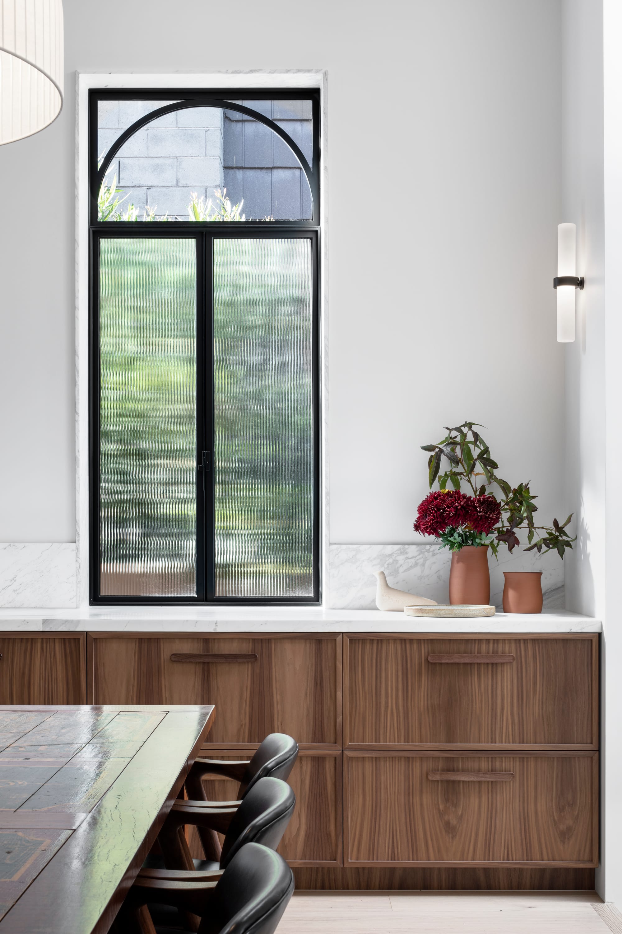 South Terrace Residence by Chan Architecture. Dining space featuring walnut wood fitted cabinet featuring white marble worktop and arched window with black steel frame.