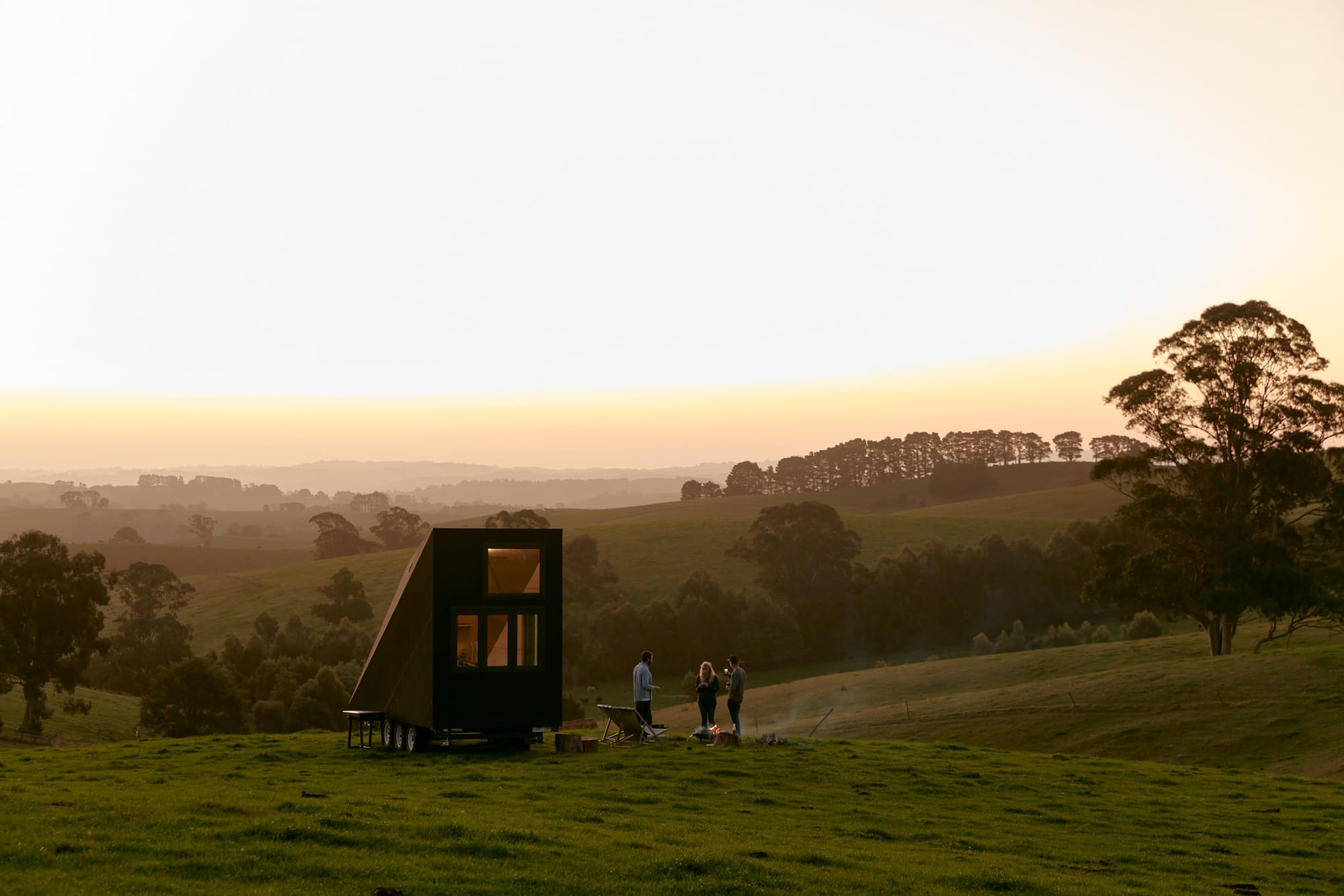 Studio Edwards by Base Cabin. Edgy shaped tiny house at sunset, standing on a trailer surrounded by nature and people barbequeing.