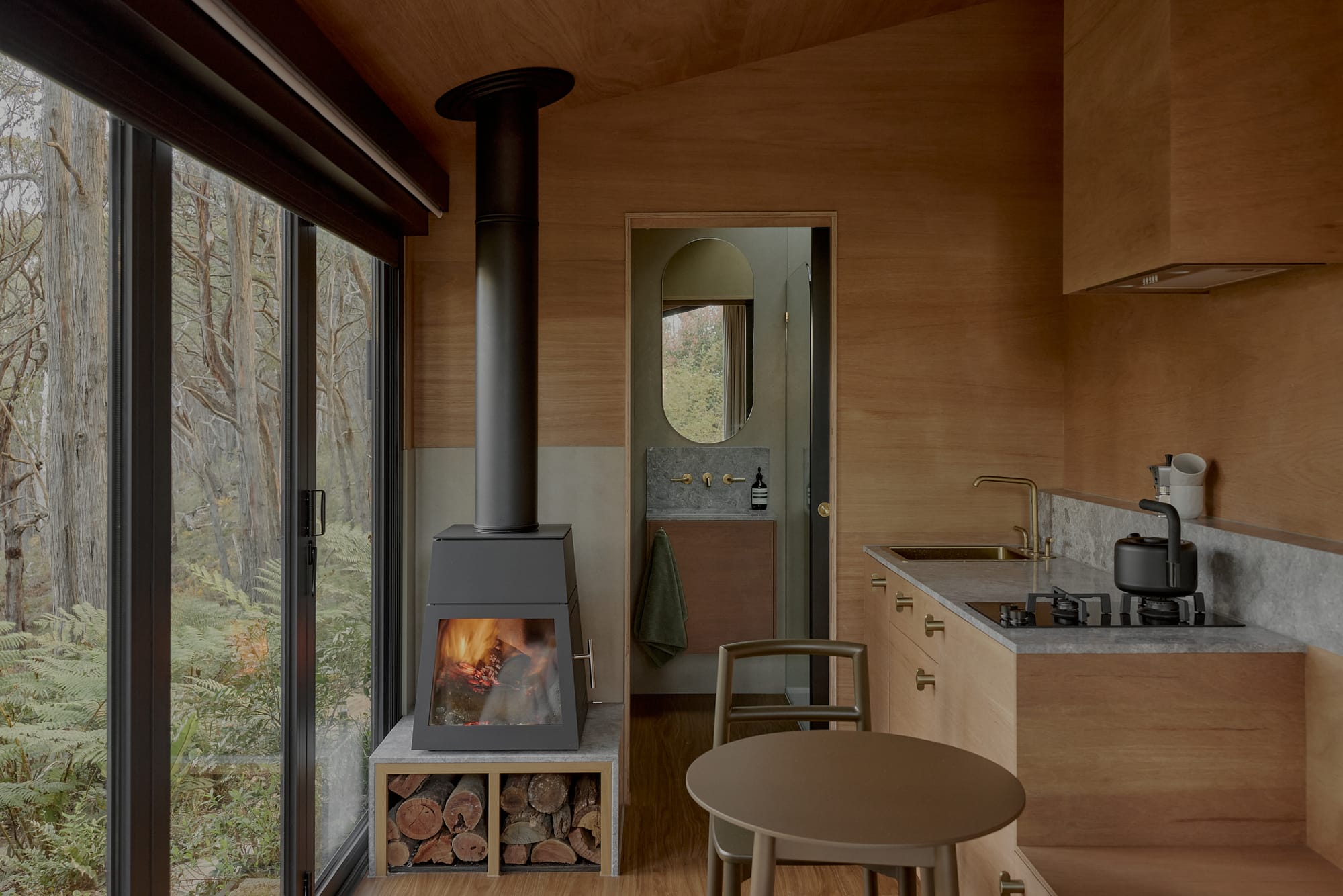 Brook James by Base Cabin. Warm and minimalistic tiny house interior with view to kitchen and bathroom featuring chimney in forest.